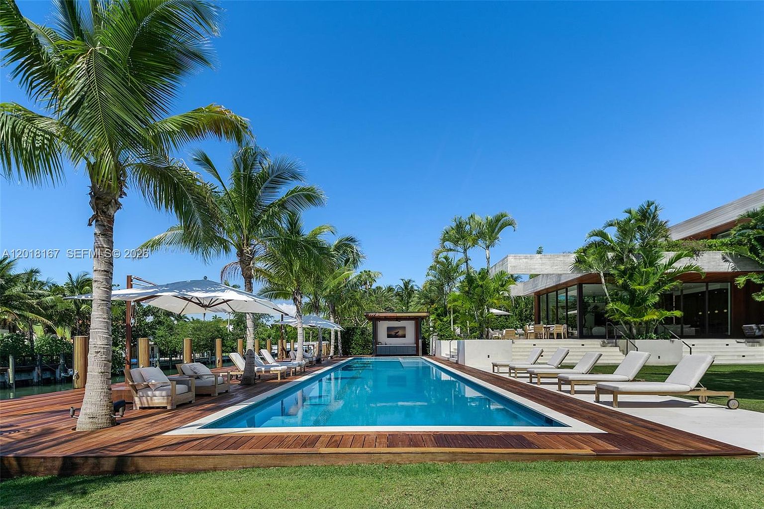 This luxurious outdoor space features a long, rectangular swimming pool set within a rich wood deck, surrounded by lush palm trees and manicured green lawn. To the right, a modern residence with large glass walls and a concrete overhang provides a sophisticated backdrop, while multiple lounge chairs offer ample seating for relaxation. The scene is captured from a low, eye-level perspective, emphasizing the seamless transition between the indoor living area and the serene waterfront pool deck.