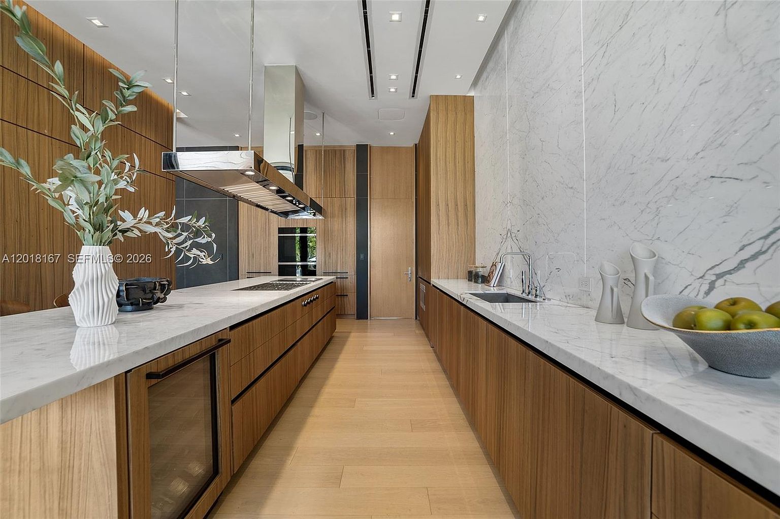 This sleek, modern kitchen features a long central island and a parallel counter, both topped with light-toned stone surfaces. The cabinetry is finished in a warm-toned wood, creating a cohesive and sophisticated aesthetic, while a large marble-patterned wall adds a touch of elegance. The perspective is a centered, eye-level shot looking down the galley-style layout, emphasizing the clean lines and high-end finishes.