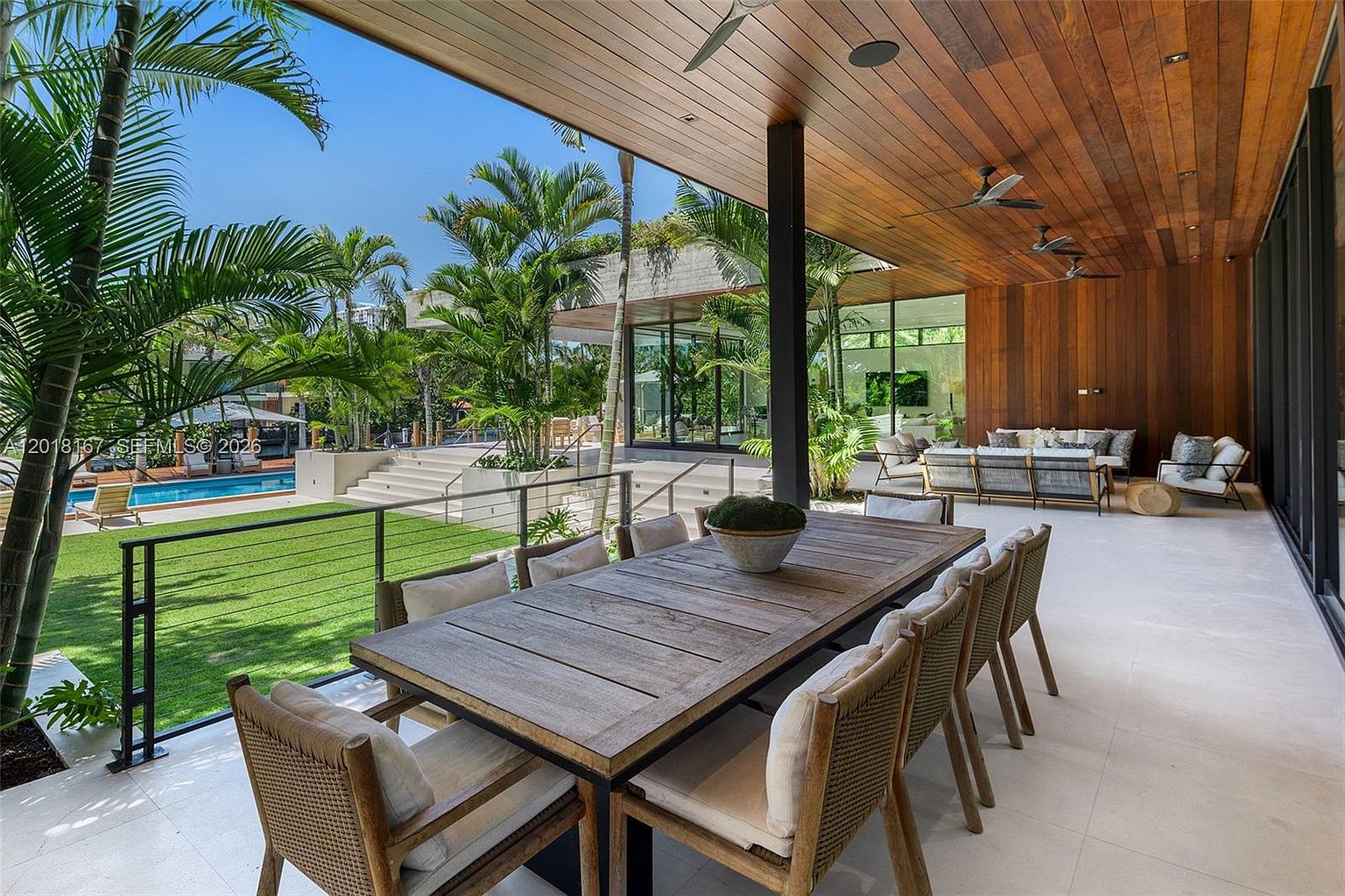 This expansive covered patio features a large wooden dining table with seating for eight, set against a backdrop of lush tropical landscaping and a swimming pool. The space is defined by a warm wood-paneled ceiling with integrated lighting and fans, transitioning seamlessly to an outdoor lounge area with comfortable seating. The perspective captures the transition from the shaded, sophisticated dining area to the bright, open lawn and pool deck, emphasizing a luxurious indoor-outdoor lifestyle.