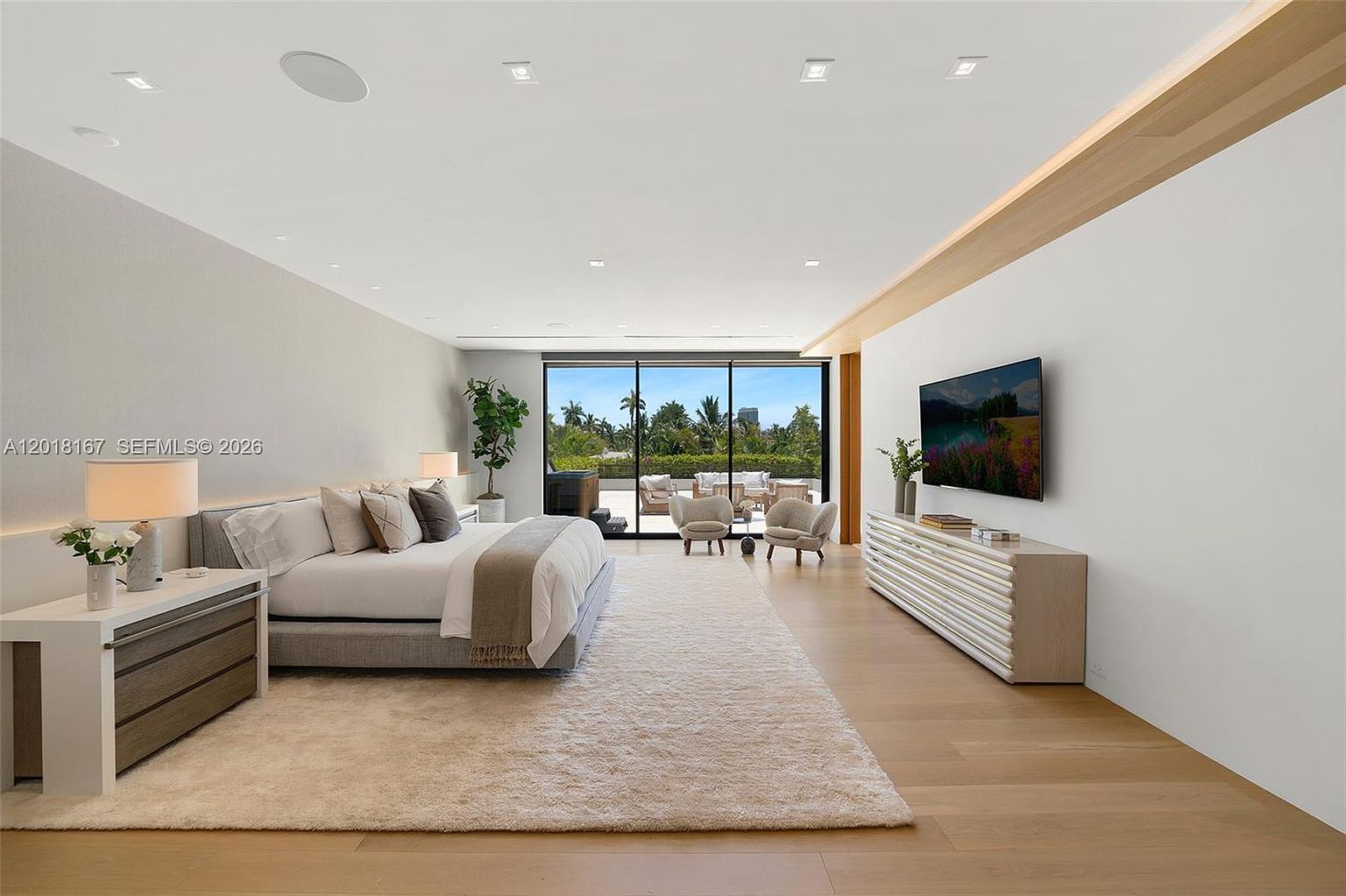 This spacious primary bedroom features light wood flooring, a neutral color palette, and a large bed centered against a wall with a minimalist bedside table and lamp. A long, textured console table sits beneath a wall-mounted television, while floor-to-ceiling glass sliding doors open to a private balcony with outdoor seating. The room is illuminated by recessed lighting and offers a bright, airy atmosphere with a view of lush greenery outside.