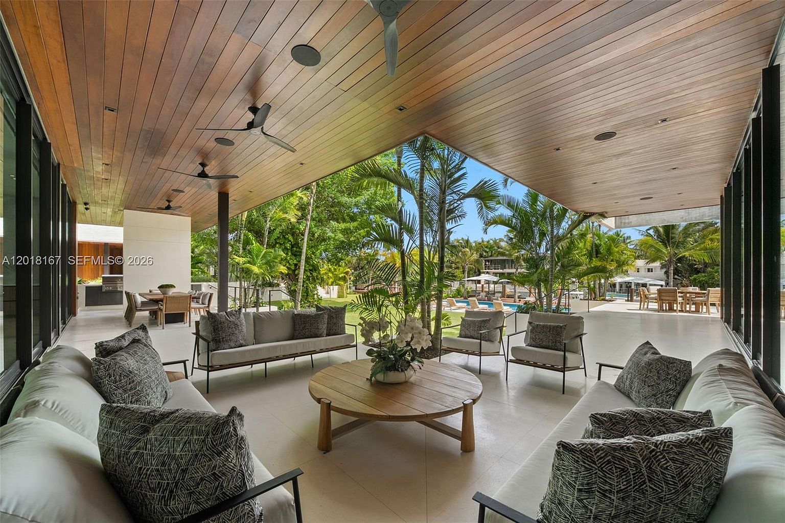 This expansive covered patio offers a sophisticated outdoor living space with a warm wood-paneled ceiling and multiple ceiling fans. The area is furnished with two plush sofas and two armchairs arranged around a central round wooden coffee table, overlooking a lush backyard with palm trees and a swimming pool. The open-air design seamlessly blends indoor comfort with the tropical landscape, creating an inviting atmosphere for relaxation or entertaining.