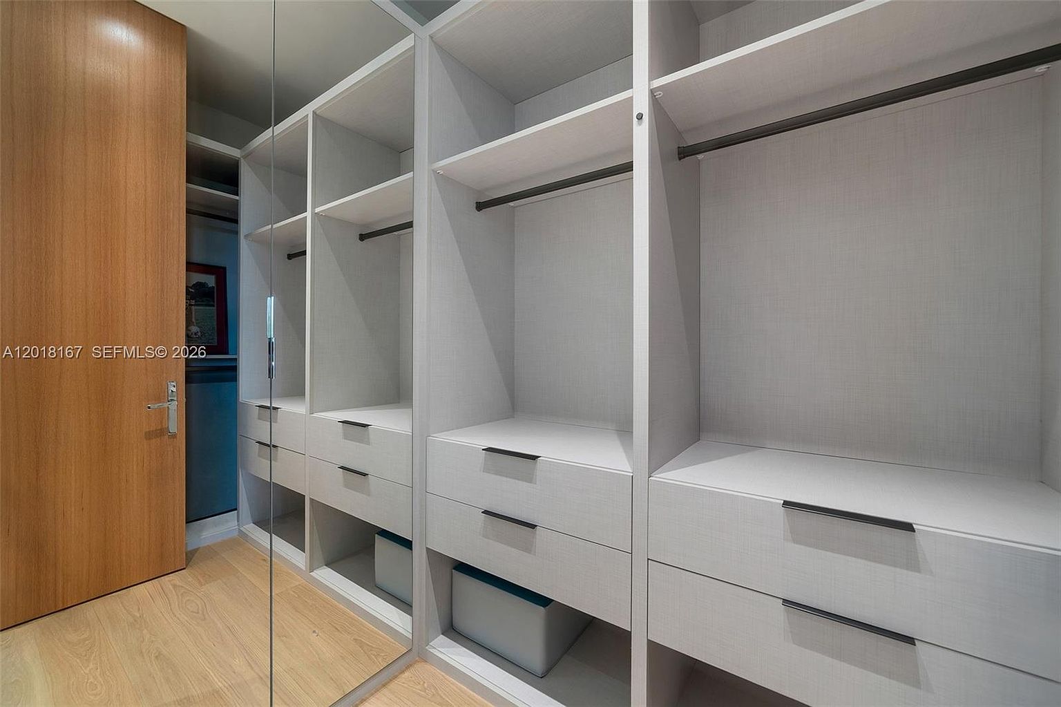 This walk-in closet features custom light-toned cabinetry with a mix of open shelving, hanging rods, and deep drawers. A large, full-height mirror panel on the left wall adds depth to the space, while light wood-look flooring provides a clean, modern aesthetic. The organized layout offers ample storage and a bright, functional atmosphere.