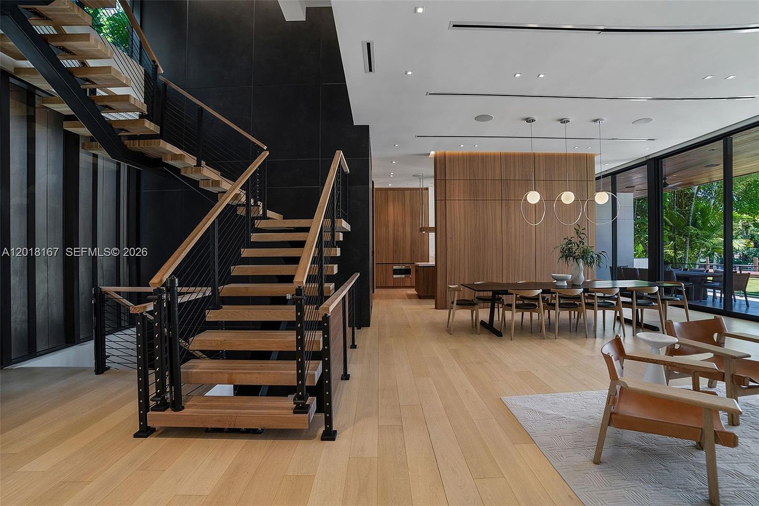 This open-concept interior space features a striking staircase with light wood treads and a black metal railing, set against a dramatic dark accent wall. To the right, a dining area is illuminated by three spherical pendant lights, with large glass walls providing a seamless view of the lush outdoor greenery. The light wood flooring and warm wood-paneled wall accents create a sophisticated, modern aesthetic that blends indoor and outdoor living.