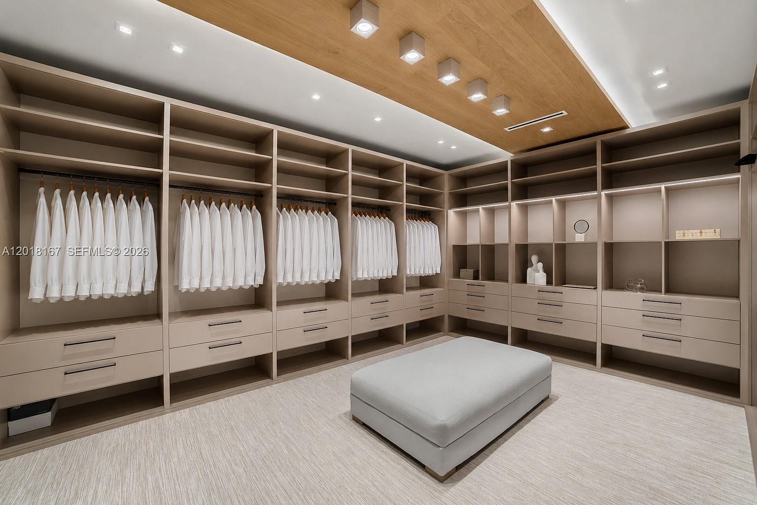This expansive, modern walk-in closet features custom light wood cabinetry with extensive hanging space and integrated drawer storage. A central upholstered ottoman provides a functional focal point, while recessed lighting and a wood-paneled ceiling detail add a sophisticated, warm aesthetic. The neutral color palette and clean lines create a luxurious and organized atmosphere.