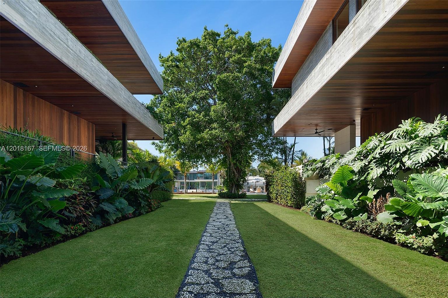 This perspective captures a lush, manicured garden path framed by two modern architectural structures with prominent wood-paneled overhangs. A central stone walkway leads toward a large, mature tree that serves as a focal point, flanked by dense tropical foliage and vibrant green lawn. The composition emphasizes a symmetrical, serene outdoor space that blends contemporary design with natural landscaping.