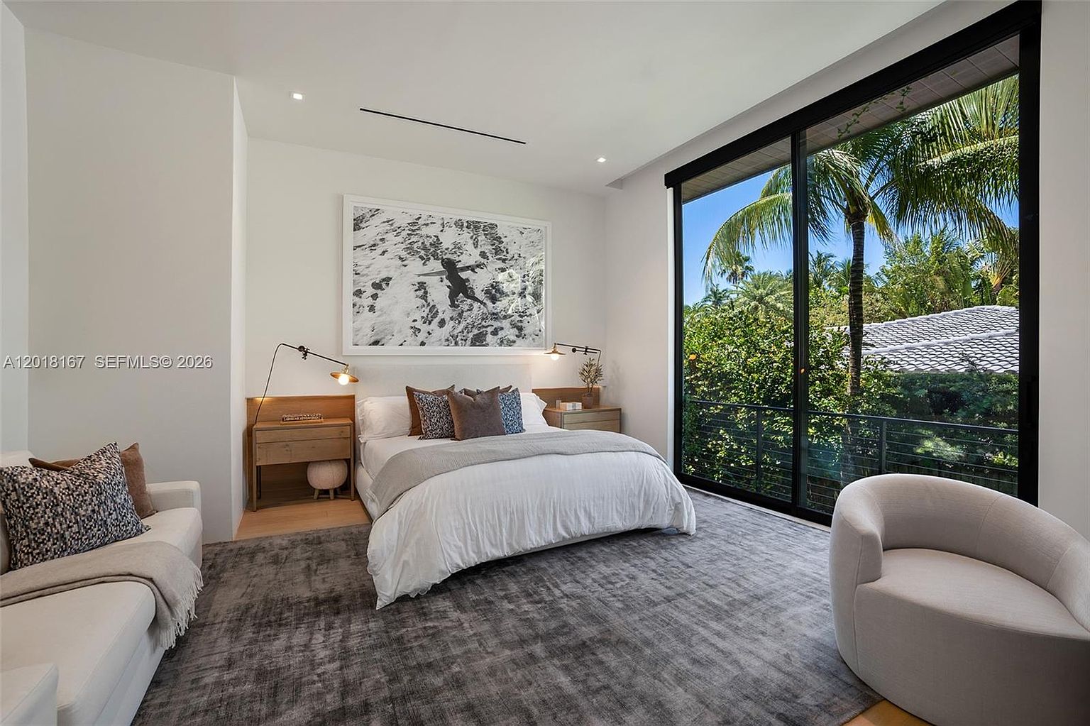 This modern primary bedroom features a neutral color palette with a large bed centered against a wall adorned with a black-and-white surf-themed art piece. To the right, expansive floor-to-ceiling windows provide a lush, green view of the surrounding trees and foliage, while a plush armchair sits in the foreground. The room is completed by a large, textured area rug, light wood nightstands with minimalist lamps, and a small sofa on the left, creating a serene and sophisticated atmosphere.