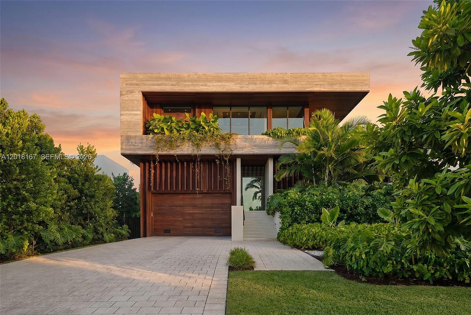 This modern residence features a striking architectural design with a prominent concrete upper level cantilevered over a warm-toned wood-paneled garage and entryway. Lush tropical landscaping surrounds the property, softening the clean lines of the structure against a vibrant sunset sky. A paved driveway leads to a set of stairs that provide access to the main entrance, creating an inviting and sophisticated curb appeal.
