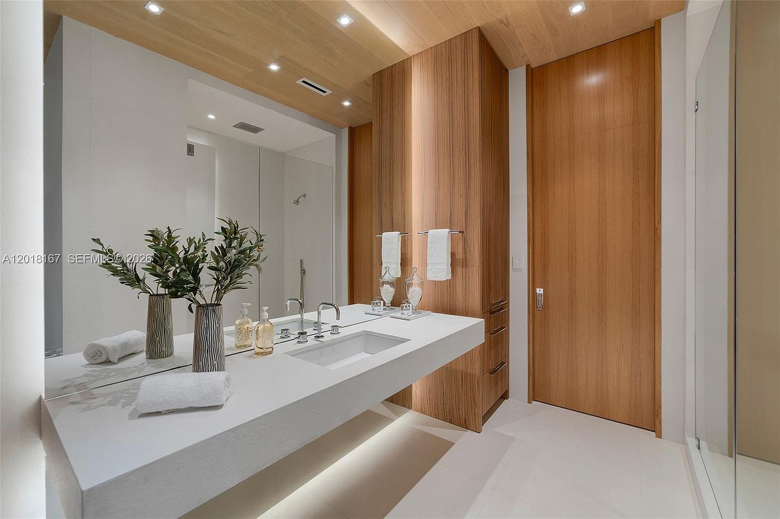 This modern bathroom features a sleek, floating vanity with a white countertop and integrated sink, set against a large mirror that enhances the sense of space. The room is accented with warm-toned wood paneling on the ceiling and a tall storage cabinet, creating a clean, minimalist aesthetic. Soft under-cabinet lighting and recessed ceiling lights provide a bright, inviting atmosphere.