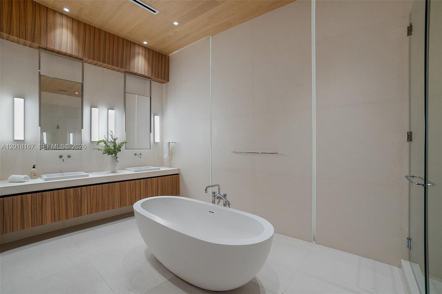 This modern primary bathroom features a sleek, freestanding white soaking tub as the central focal point. A long, wall-mounted vanity with light wood cabinetry and dual vessel sinks sits beneath large, rectangular mirrors and integrated vertical lighting. The room is finished with neutral, large-format wall tiles and a warm wood-paneled ceiling, creating a clean and sophisticated spa-like atmosphere.
