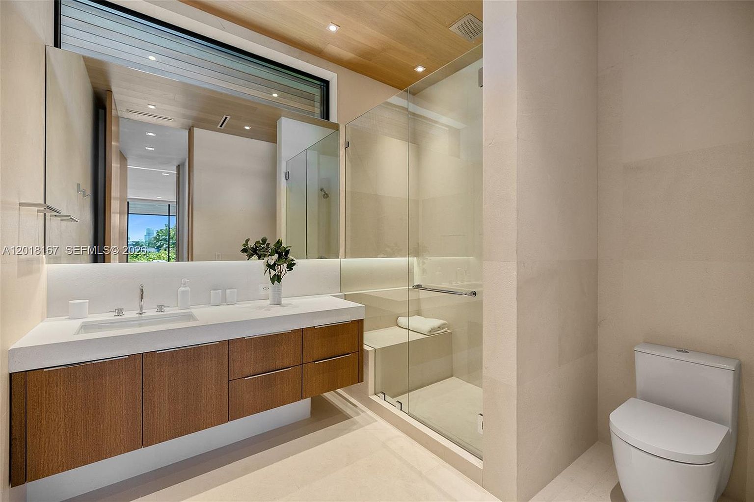 This modern bathroom features a floating vanity with light wood cabinetry and a sleek, integrated sink. A large, frameless glass shower enclosure includes a built-in bench and a white towel, while a minimalist white toilet sits to the right. The space is finished with neutral-toned walls and a warm wood-paneled ceiling, creating a clean and contemporary aesthetic.