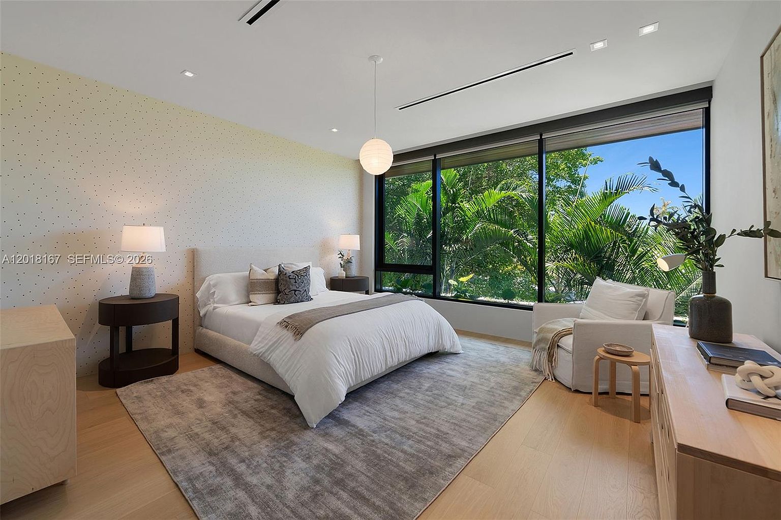 This bright and airy primary bedroom features a neutral color palette with a textured, patterned accent wall behind the bed. A large, floor-to-ceiling window wall provides an expansive view of lush tropical greenery outside, filling the room with natural light. The space is furnished with a plush bed, two bedside tables with lamps, and a comfortable armchair, all set atop a large area rug over light wood flooring.