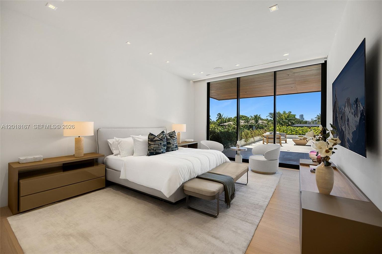 This spacious primary bedroom features a neutral color palette and a minimalist aesthetic, centered around a large bed with patterned accent pillows. The room is anchored by a light-toned area rug over wood flooring and includes a sleek bench at the foot of the bed, two bedside tables with lamps, and a low-profile media console. Large floor-to-ceiling windows provide an expansive view of the outdoor patio and lush greenery, creating a seamless transition between indoor and outdoor living spaces.