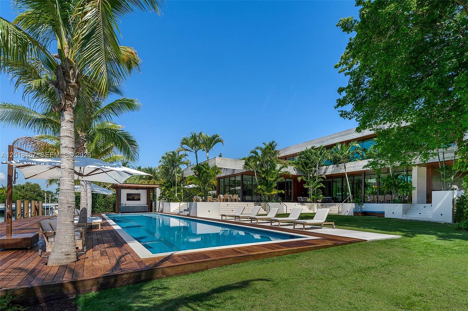 This expansive rear exterior view showcases a modern, multi-level residence with a large rectangular swimming pool as the central feature. The pool is surrounded by a rich wood deck and a lush green lawn, complemented by several lounge chairs and a shaded cabana area. The architecture features clean lines, large glass walls, and a contemporary aesthetic that seamlessly integrates the indoor and outdoor living spaces.