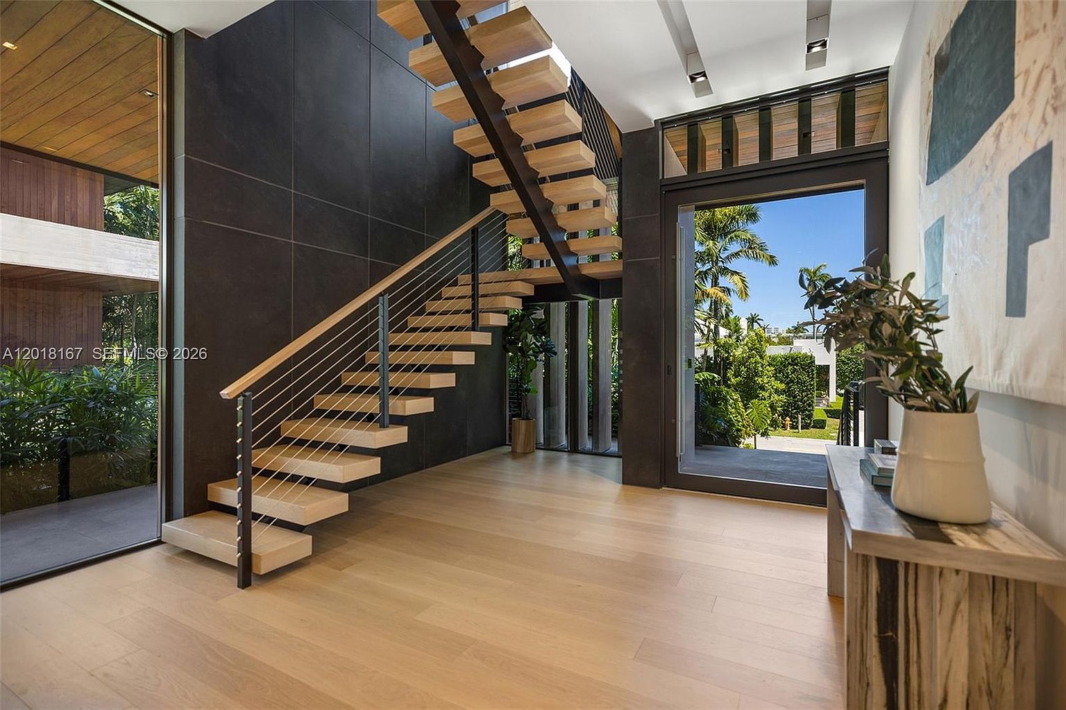 This modern entryway features a striking floating staircase with light wood treads and a sleek metal cable railing. The space is anchored by a dark, large-format tile feature wall that contrasts with the light wood flooring and bright, airy atmosphere. A large glass door provides a seamless transition to the lush outdoor greenery, creating a sophisticated and open architectural aesthetic.