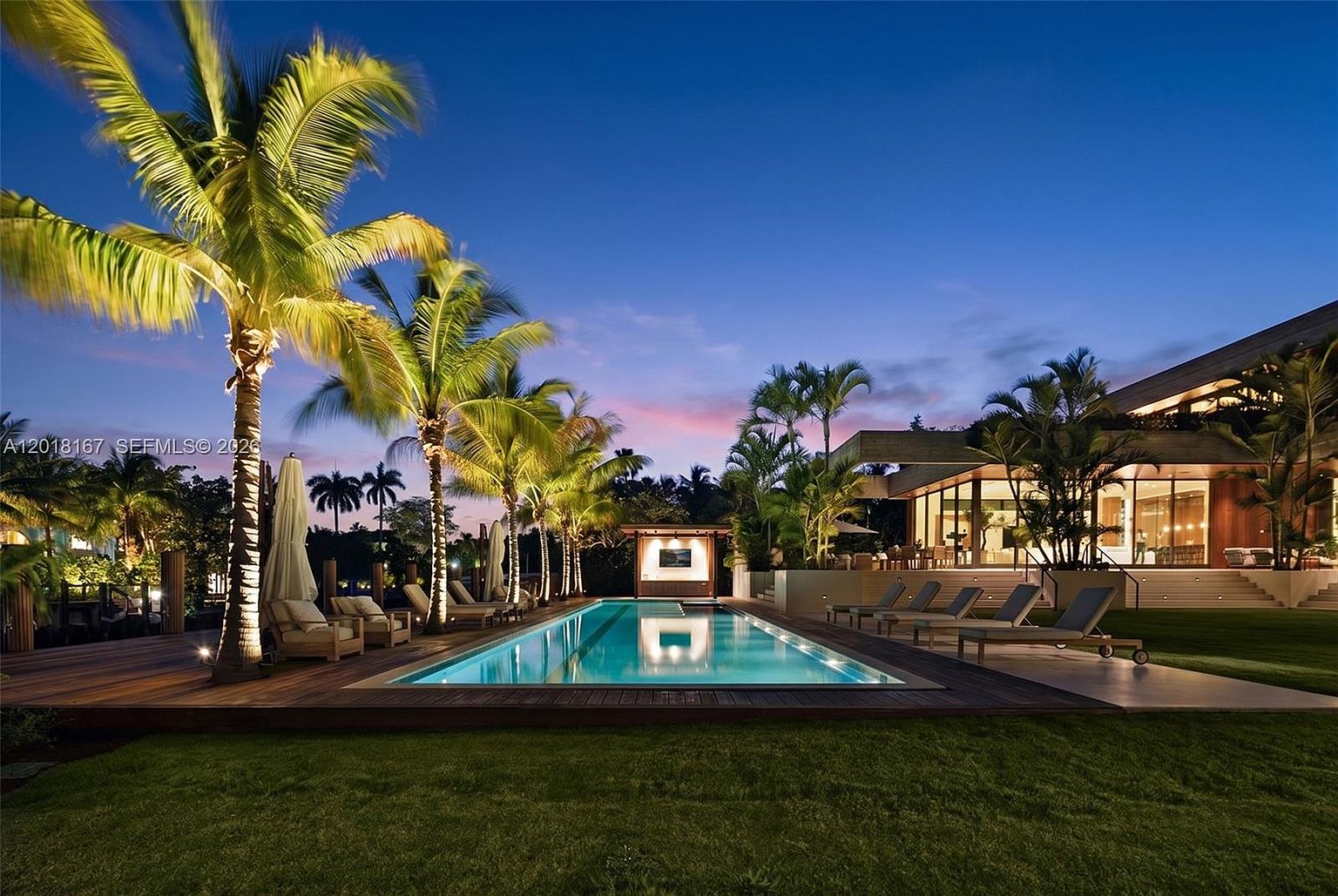 This stunning twilight view captures a luxurious backyard oasis featuring a long, rectangular swimming pool as the centerpiece. The pool is surrounded by a dark wood deck, manicured lawn, and lush palm trees, with a modern residence featuring large glass walls visible in the background. The scene is illuminated by warm landscape lighting, creating an inviting and sophisticated atmosphere perfect for evening relaxation.