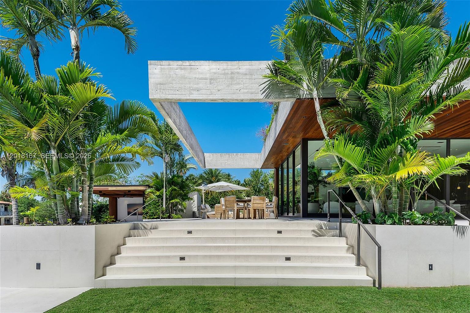 This modern outdoor living space features a wide set of concrete steps leading up to a dining area shaded by a large, minimalist concrete frame. The architecture emphasizes clean lines and natural textures, with lush palm trees framing the view and large glass walls connecting the interior to the patio. The scene captures a bright, sunny day, highlighting the seamless transition between the manicured lawn and the sophisticated outdoor entertaining area.
