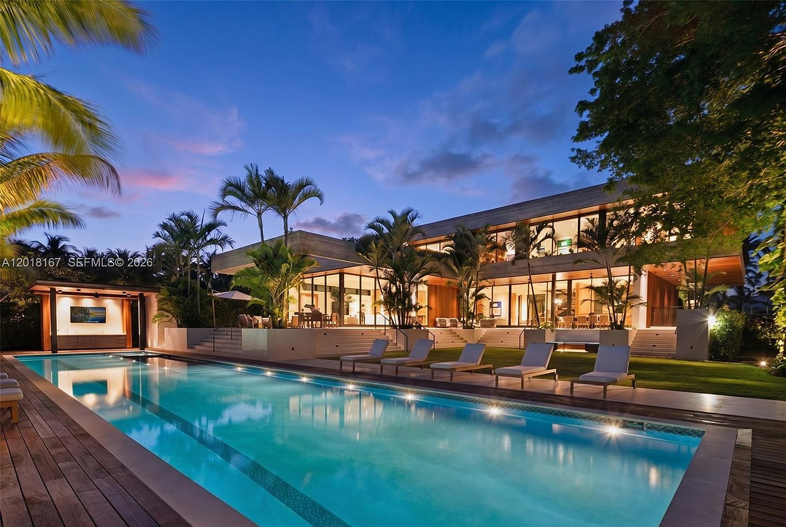 This stunning contemporary residence is captured at dusk, showcasing a sprawling multi-level backyard centered around a large, illuminated rectangular swimming pool. The home features extensive glass walls that provide a seamless transition between the interior living spaces and the outdoor patio, which is furnished with multiple lounge chairs. Lush tropical landscaping and mature trees frame the property, creating a serene and luxurious atmosphere.