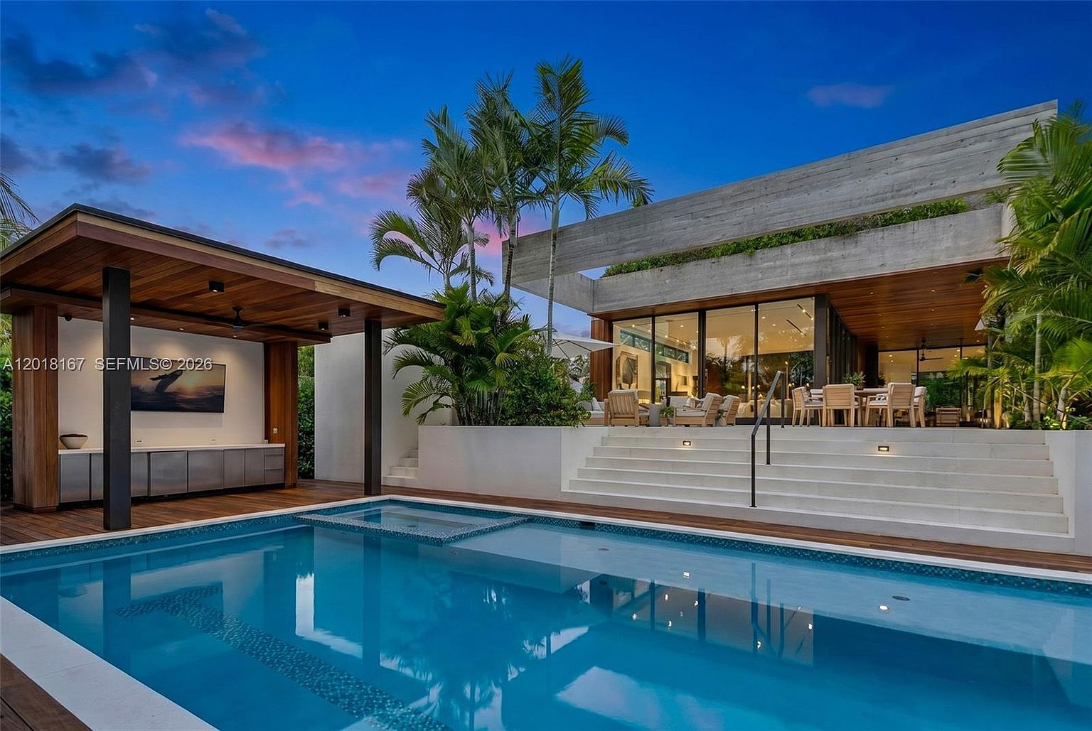 This luxurious outdoor living space features a pristine swimming pool with an integrated spa, set against a modern architectural backdrop. A covered patio area includes a built-in outdoor kitchen and a wall-mounted television, while wide concrete steps lead up to a spacious terrace with dining furniture and large glass walls. The scene is captured at dusk, highlighting the warm interior lighting and the sleek, contemporary design of the residence.