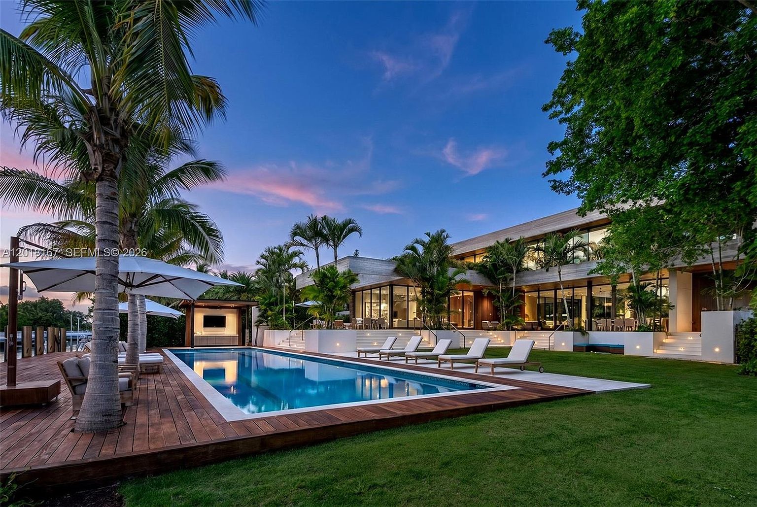 This stunning rear exterior view showcases a modern luxury residence featuring expansive glass walls and a sleek, contemporary architectural design. A rectangular swimming pool serves as the focal point, surrounded by a rich wood deck and a manicured lawn, with several lounge chairs positioned for relaxation. The scene is set during a vibrant sunset, with palm trees framing the property and soft landscape lighting illuminating the tiered patio levels.