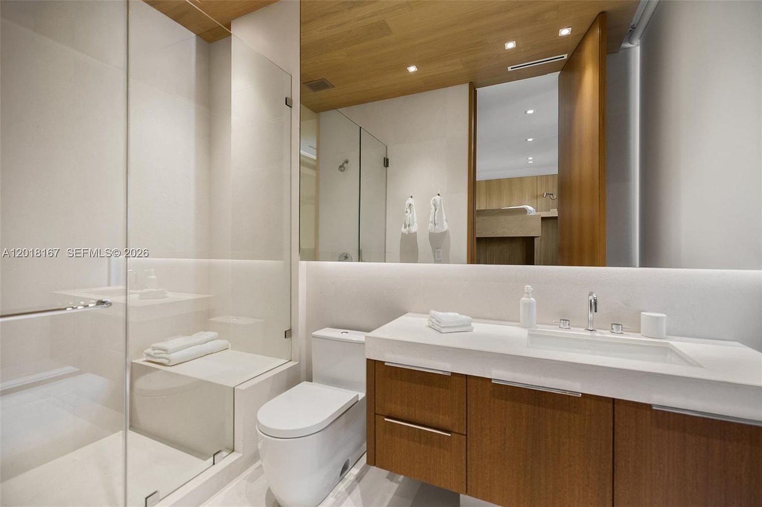 This modern bathroom features a sleek vanity with a wood-toned cabinet and a white countertop, complemented by a large mirror that reflects the warm wood-paneled ceiling. A glass-enclosed shower stall with a built-in bench sits adjacent to a contemporary toilet, creating a clean and minimalist aesthetic. The space is illuminated by recessed lighting, providing a bright and sophisticated atmosphere.