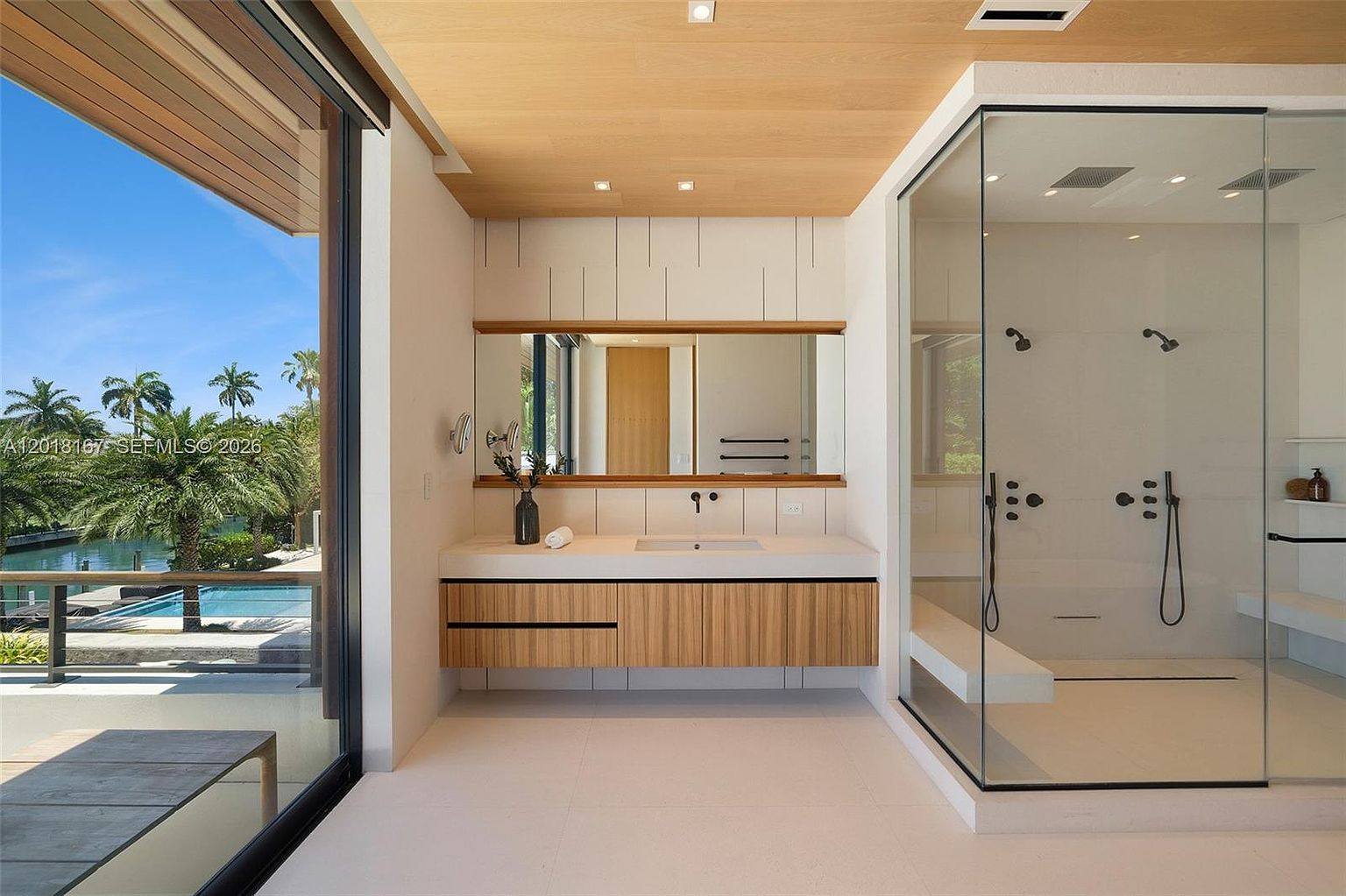 This modern primary bathroom features a floating light wood vanity with a sleek countertop and a wide mirror set against a paneled wall. To the right, a spacious glass-enclosed walk-in shower includes dual shower heads and a built-in bench. The room is bathed in natural light from large windows that offer a serene view of an outdoor pool and lush tropical landscaping.
