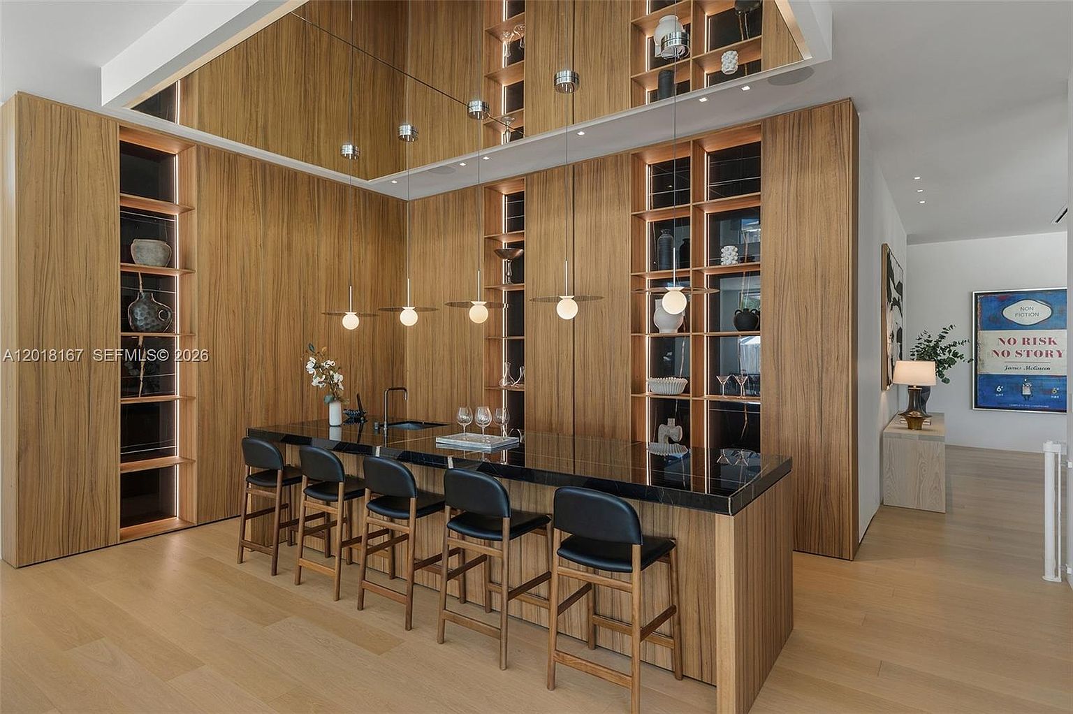 This sophisticated kitchen features a large, dark stone-topped island with seating for five, set against a backdrop of warm-toned wood cabinetry and integrated shelving. The space is accented by a mirrored ceiling detail and modern pendant lighting, creating a sleek, high-end aesthetic. A piece of framed wall art adds a touch of personality to the adjacent hallway area, completing the contemporary design.