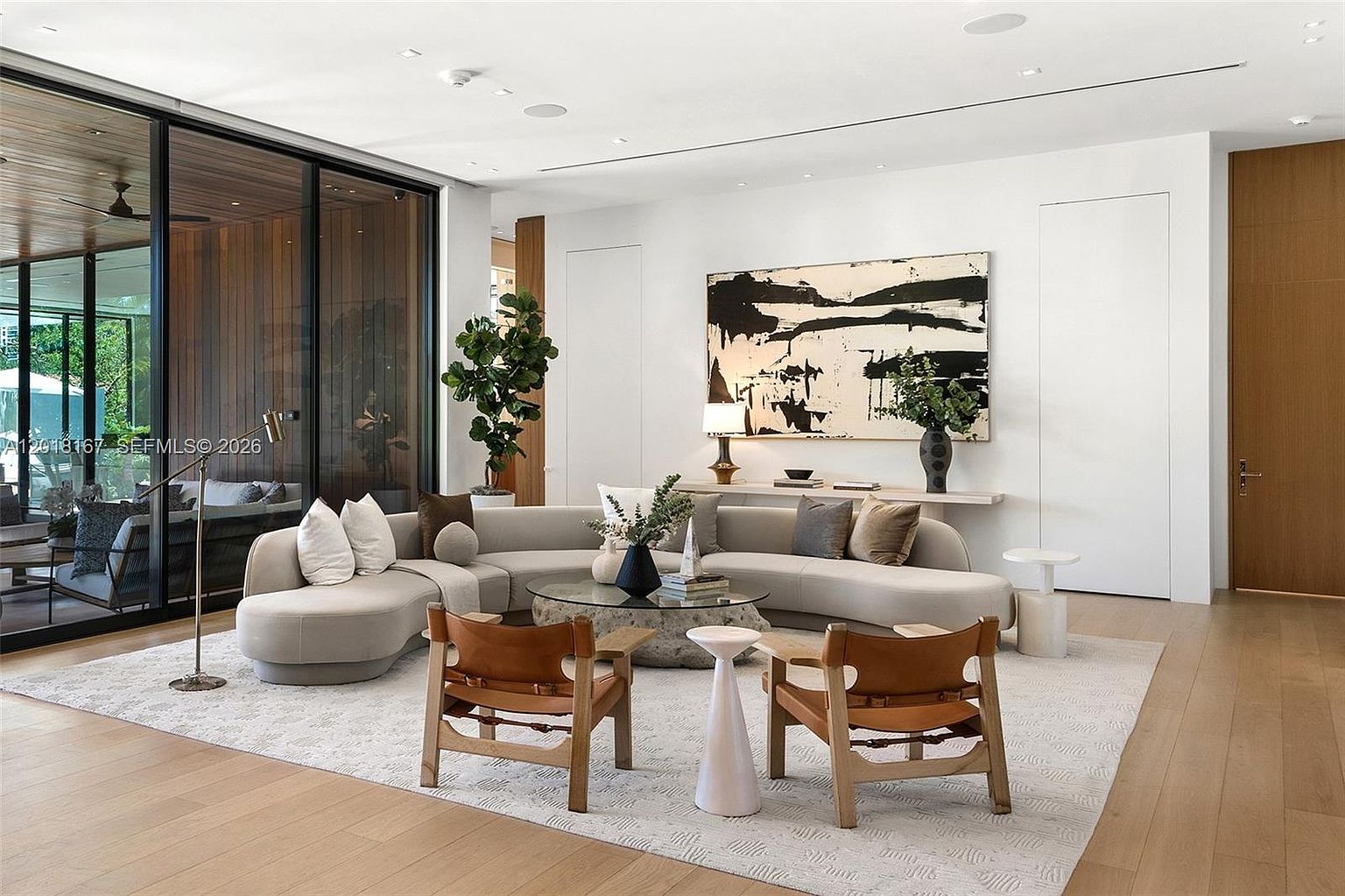 This sophisticated living room features a large, curved sectional sofa arranged around a unique, organic-shaped stone coffee table. The space is anchored by two leather-accented wooden chairs and a neutral area rug, set against a backdrop of light wood flooring and crisp white walls. Large floor-to-ceiling glass panels provide a seamless transition to an outdoor patio, while a bold abstract painting serves as a striking focal point above a minimalist console table.
