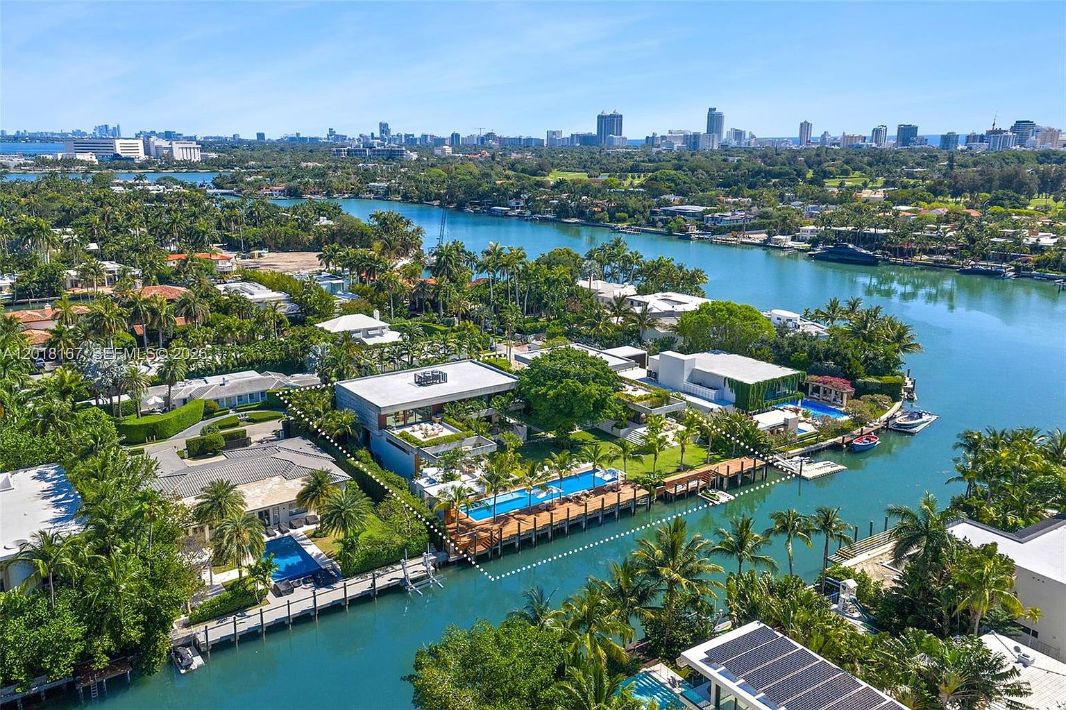 This high-angle aerial view captures a luxurious waterfront estate featuring a modern architectural design with flat rooflines and expansive outdoor living spaces. The property includes a private swimming pool, a wooden dock extending into the canal, and lush tropical landscaping that integrates seamlessly with the surrounding coastal environment. The perspective highlights the prime location of the home, showcasing its direct water access and proximity to the broader cityscape in the distance.