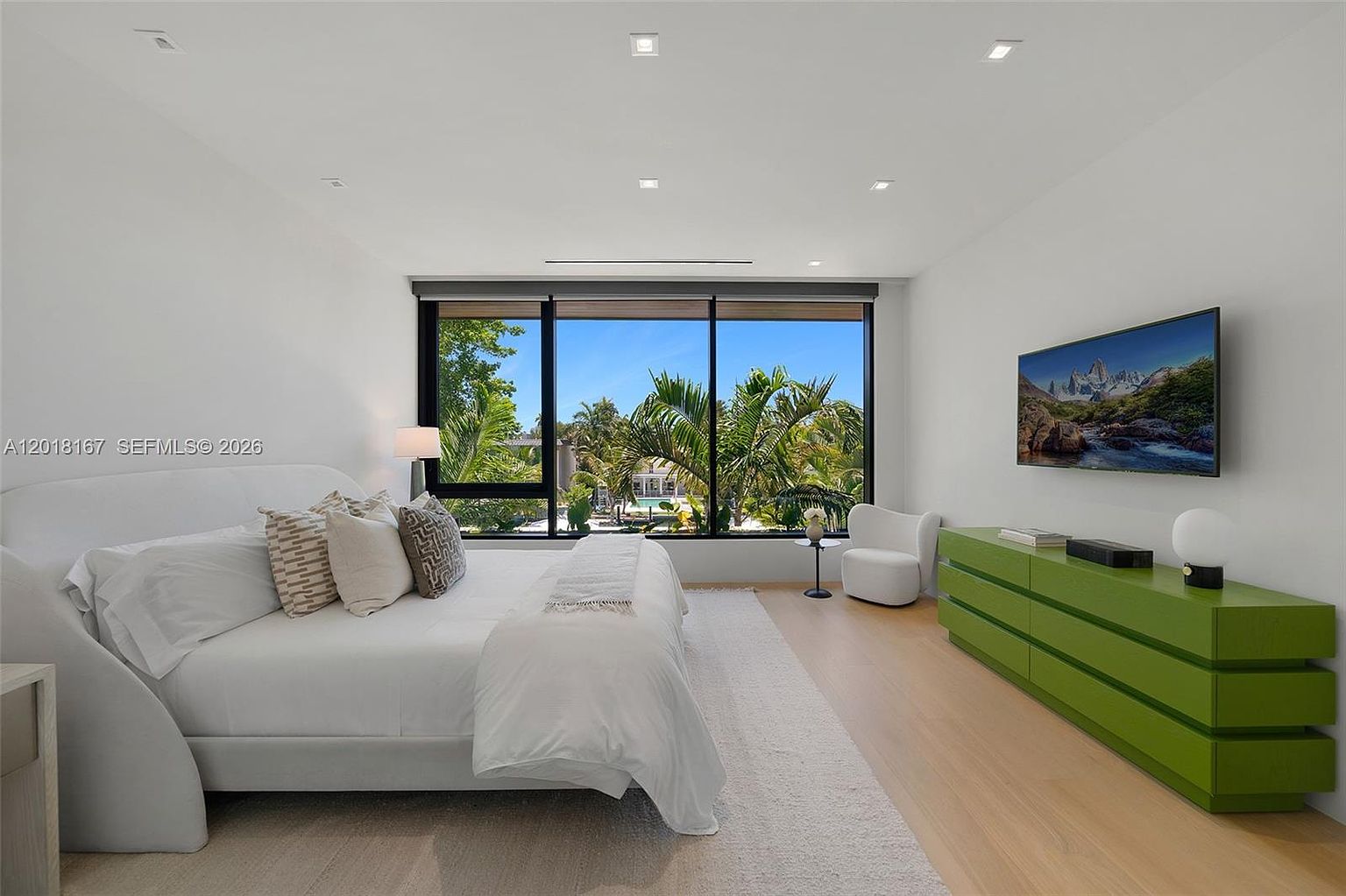 This bright and airy primary bedroom features a minimalist aesthetic with white walls, light wood flooring, and a large bed centered against the wall. A striking lime-green dresser adds a bold pop of color, while large windows provide an expansive view of the lush tropical greenery outside. The room is accented by a modern white armchair and recessed ceiling lighting, creating a serene and contemporary atmosphere.