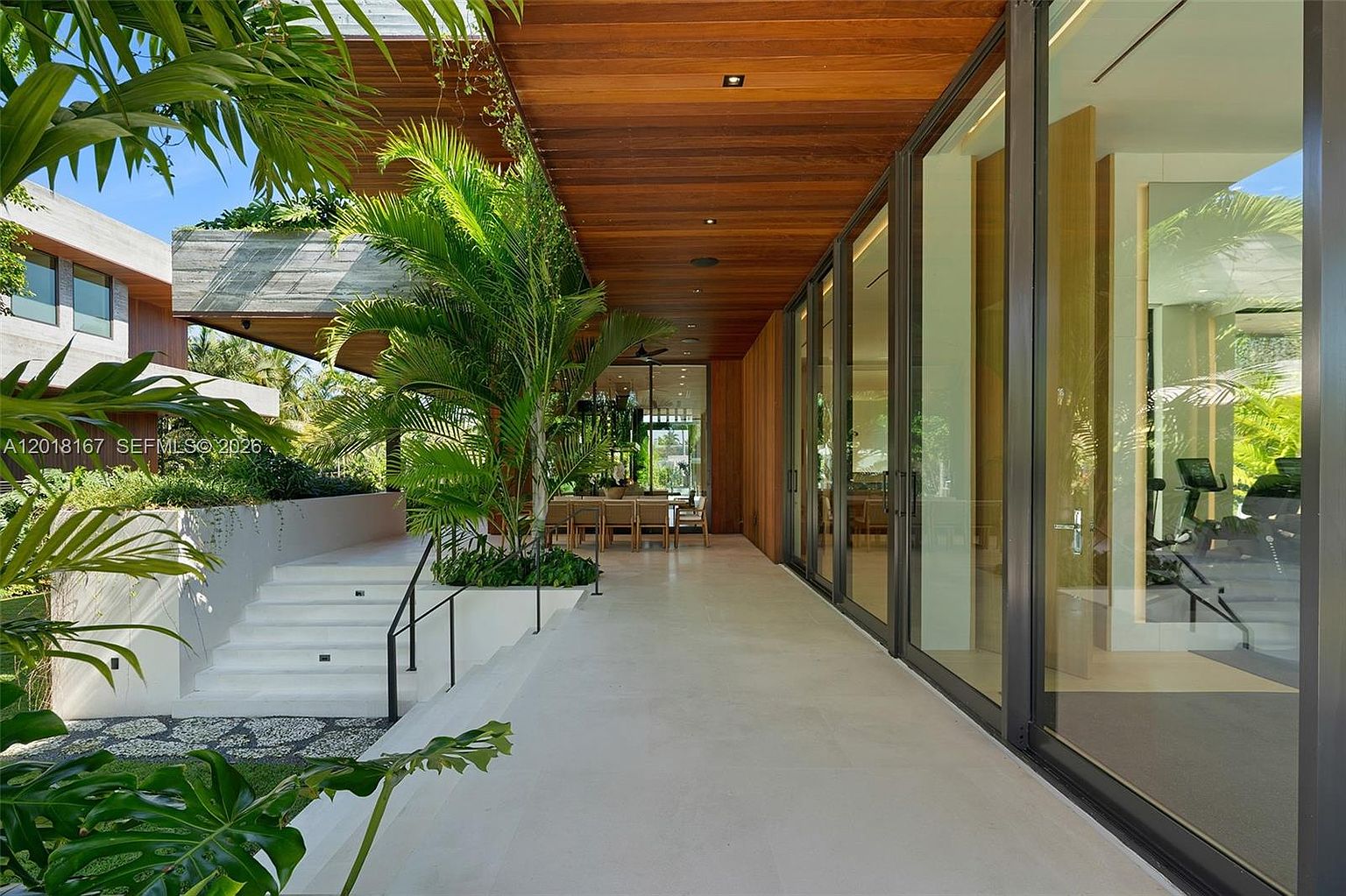 This modern covered patio features a sleek, light-toned stone floor and a warm wood-paneled ceiling that extends from the interior. Large glass panels provide a seamless transition to the indoor living spaces, while a set of white stairs leads up to an elevated garden area with lush tropical landscaping. The perspective captures the expansive, airy nature of the outdoor living space, emphasizing the integration of contemporary architecture with natural elements.