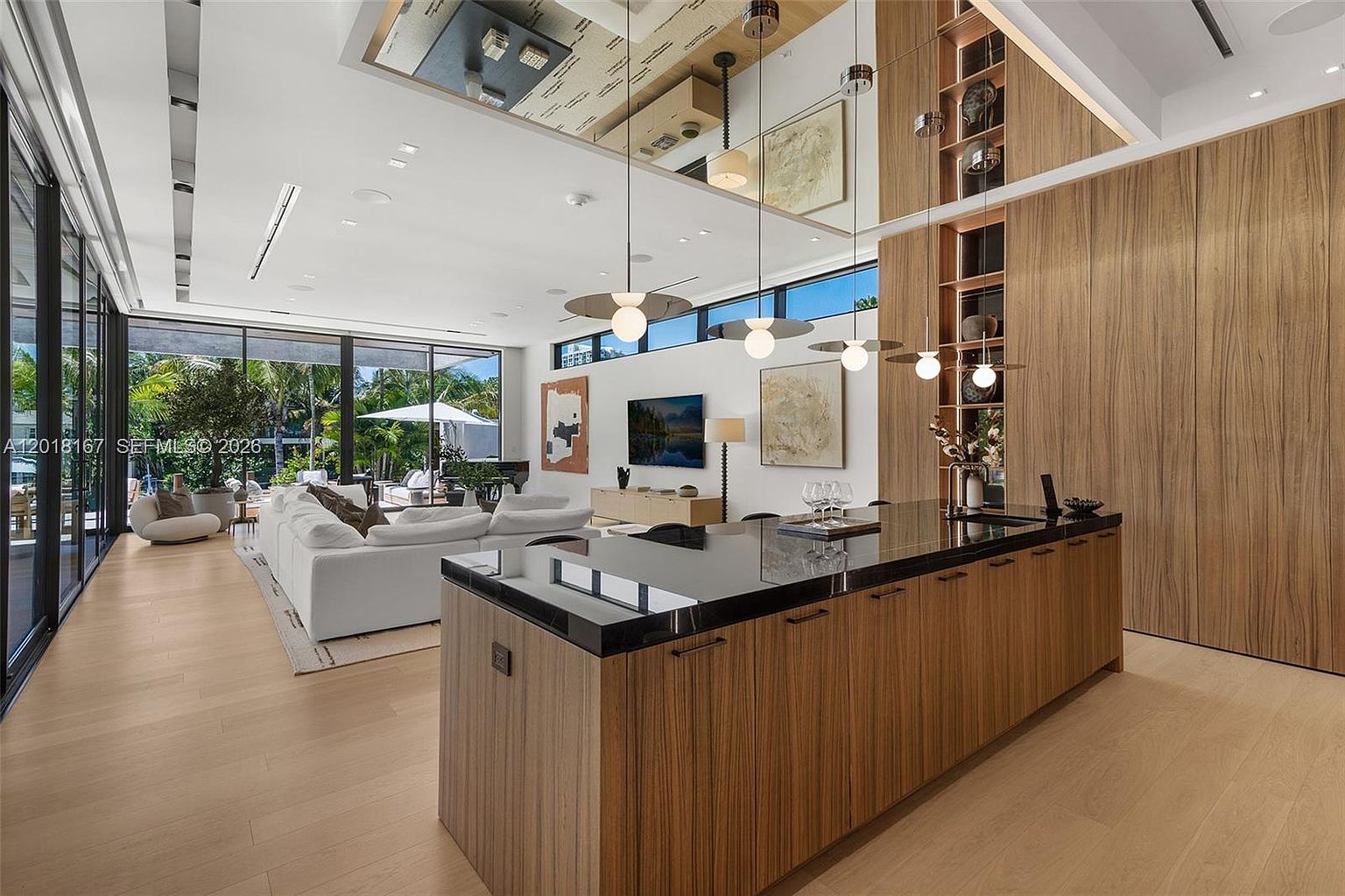 This open-concept living space features a sleek kitchen island with a dark stone countertop and light wood cabinetry, seamlessly transitioning into a bright living area. The room is defined by expansive floor-to-ceiling glass walls that provide a clear view of the lush outdoor patio and pool area. A mirrored ceiling detail above the kitchen adds a sense of height and luxury, while modern pendant lights hang over the island to create a sophisticated, contemporary atmosphere.