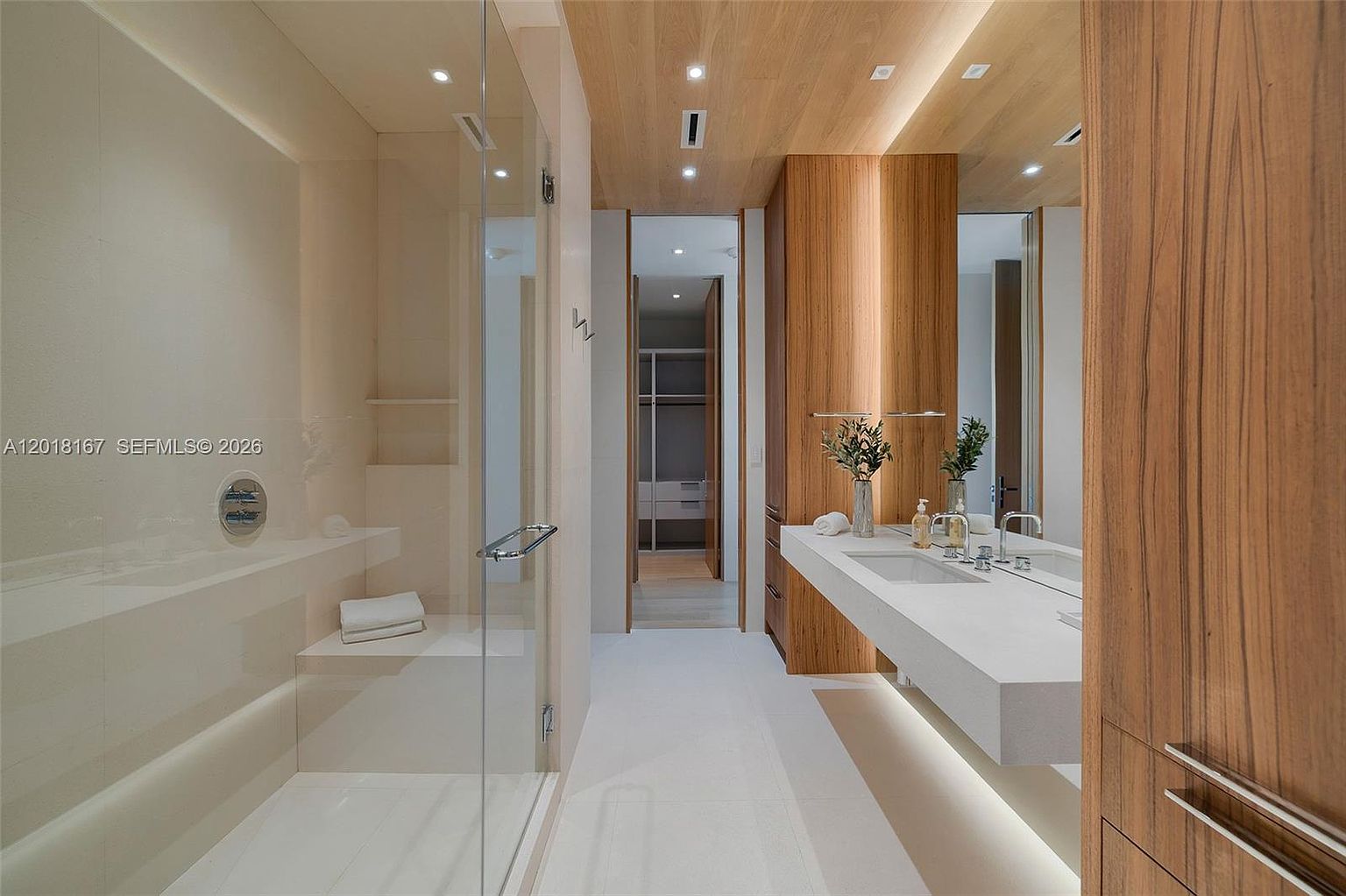 This modern primary bathroom features a sleek, minimalist design with a large walk-in shower enclosed by glass panels and a long, floating vanity with dual sinks. The space is accented by warm wood-toned ceiling and cabinetry, creating a sophisticated and spa-like atmosphere. A clear view into the adjacent walk-in closet adds to the open, airy feel of the suite.