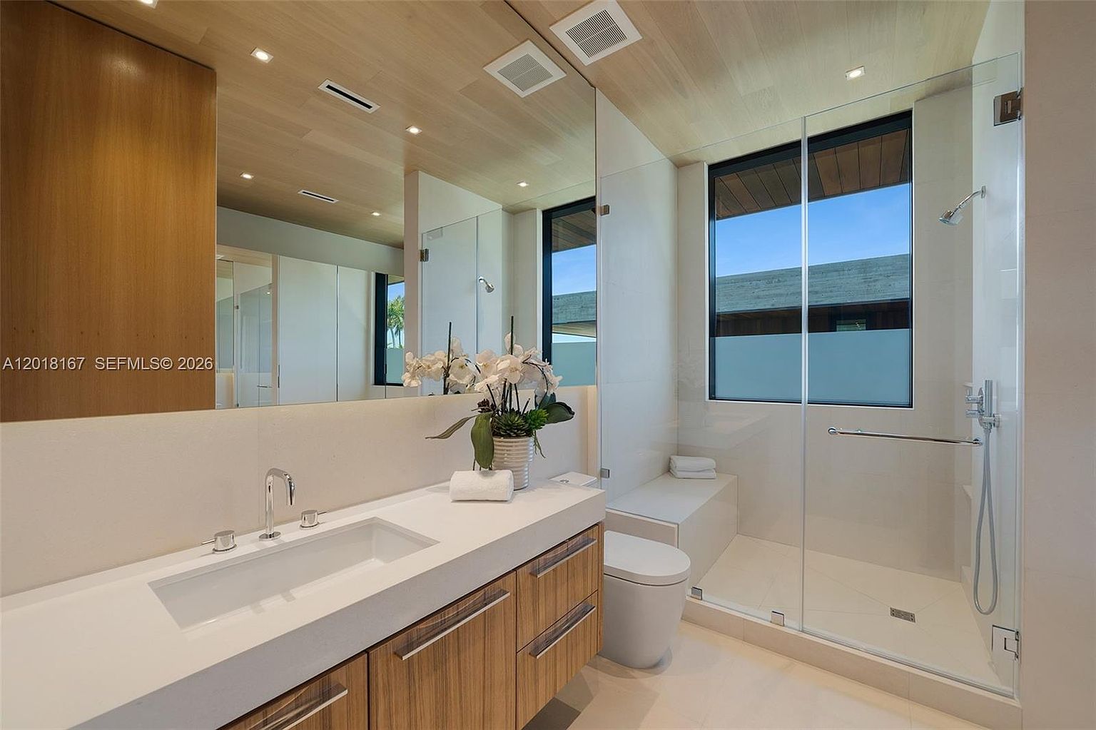 This modern bathroom features a sleek vanity with a light wood finish and a white countertop, complemented by a large wall-to-wall mirror. A glass-enclosed walk-in shower sits adjacent to a minimalist toilet and a built-in bench, all set against neutral-toned walls. The space is brightened by recessed lighting and a large window that allows natural light to fill the room.