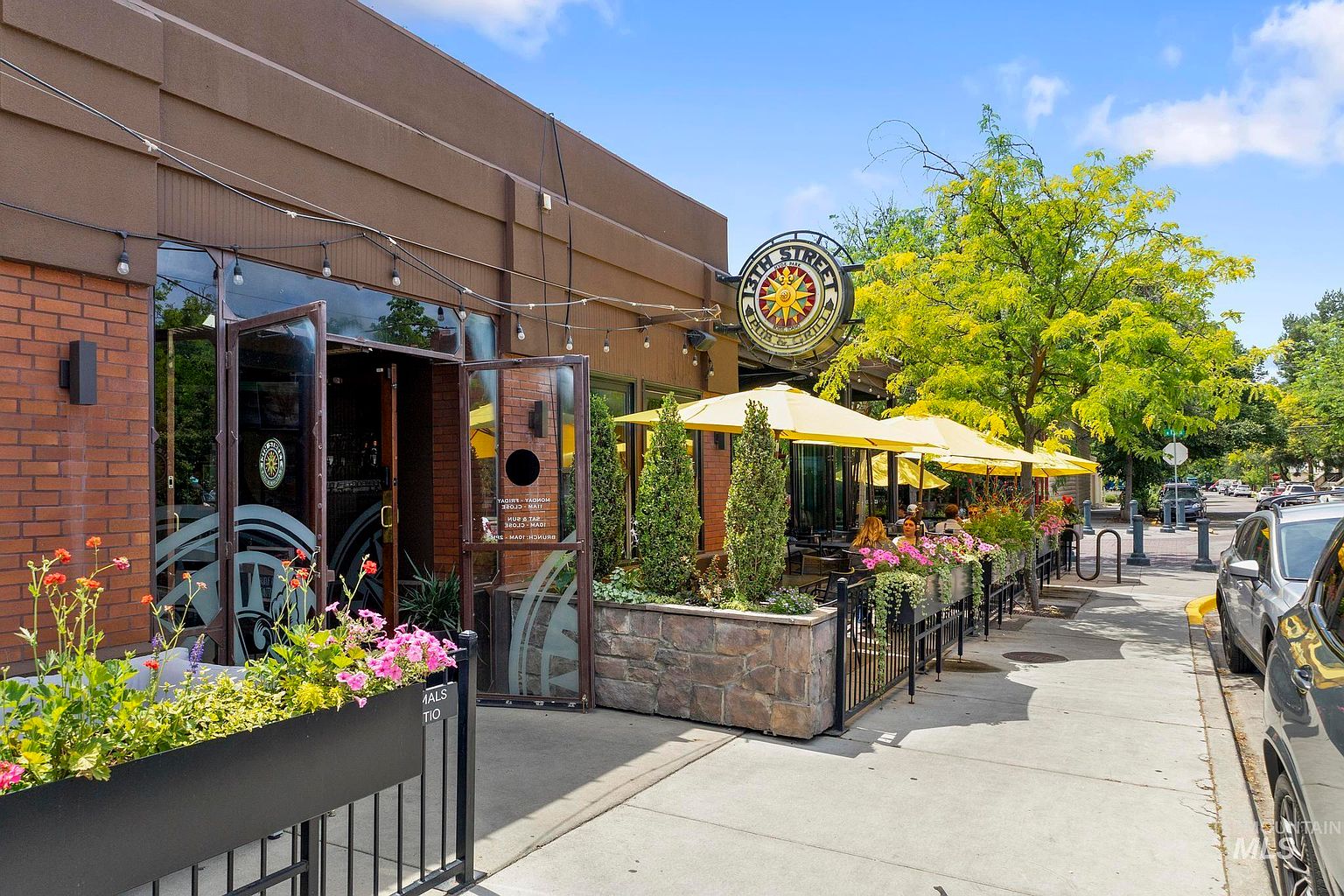 This street-level view captures the inviting facade of a commercial pub and grill, featuring a brick exterior and a prominent circular sign. A sidewalk patio area is defined by stone-clad planters and yellow umbrellas, creating a vibrant outdoor dining space. The perspective looks down the sidewalk, showcasing the restaurant's entrance and the surrounding neighborhood atmosphere on a sunny day.