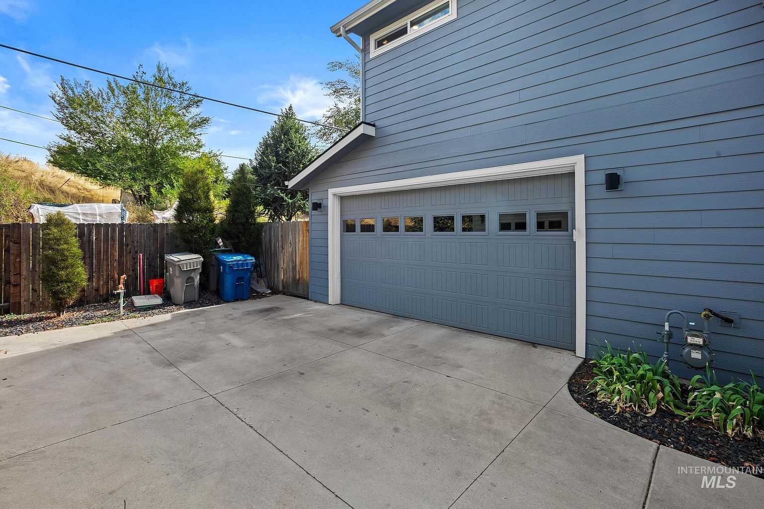 This image captures the side exterior of a home, focusing on a single-car garage with a blue-grey paneled door and a row of small windows. A concrete driveway leads up to the garage, bordered by a small garden bed with green plants and a utility meter. To the left, a wooden fence separates the property from a sloped, grassy area, with two trash bins positioned near the fence line.