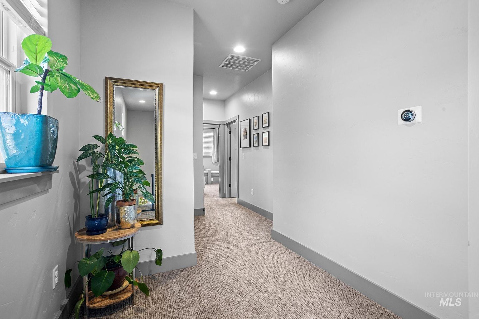 This interior hallway features neutral light-gray walls, plush carpeting, and a clean, modern aesthetic. To the left, a decorative plant sits on a tiered stand next to a tall, gold-framed mirror, while a digital thermostat is mounted on the right wall. The perspective looks down the corridor toward an open doorway, showcasing a bright and well-maintained transition space within the home.