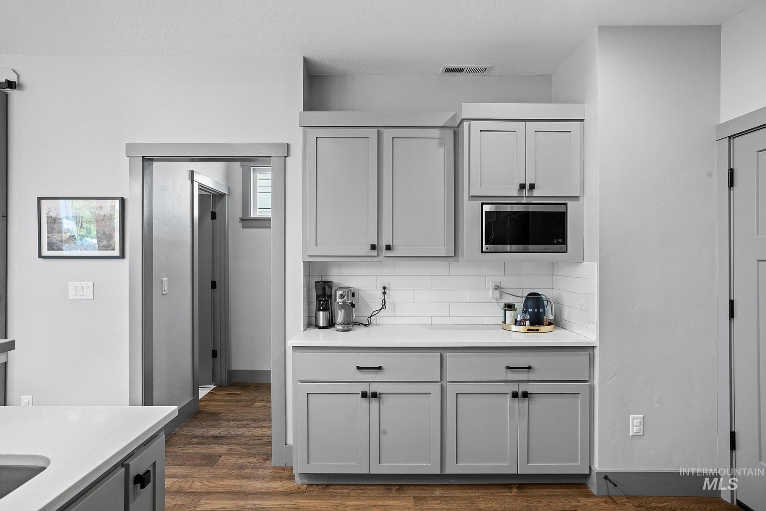 This kitchen area features a dedicated coffee station or butler's pantry with light gray cabinetry and a crisp white countertop. A white subway tile backsplash provides a clean aesthetic, complemented by a built-in microwave and a small appliance collection. The space is framed by a doorway leading to a hallway, showcasing a cohesive design with wood-look flooring and neutral wall tones.