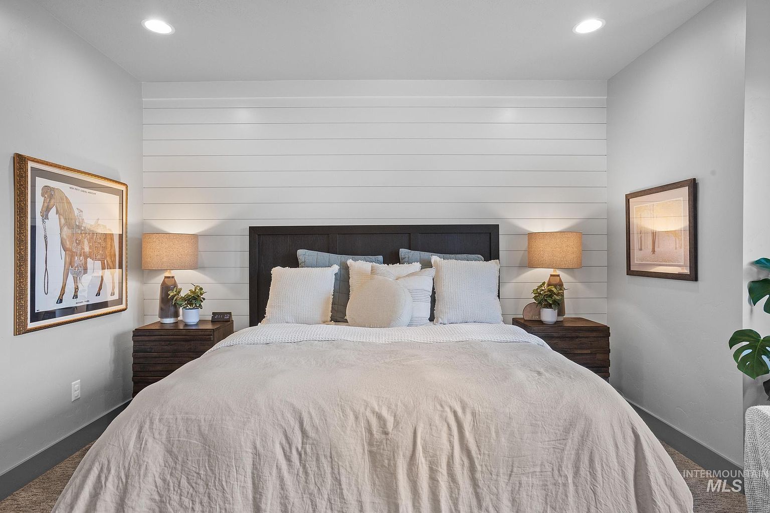 This primary bedroom features a central bed with a dark wood headboard set against a white shiplap accent wall. The room is symmetrically arranged with matching nightstands, table lamps, and framed artwork on either side, creating a balanced and serene atmosphere. A neutral-toned comforter and layered pillows add texture, while recessed lighting provides a soft, warm glow throughout the space.