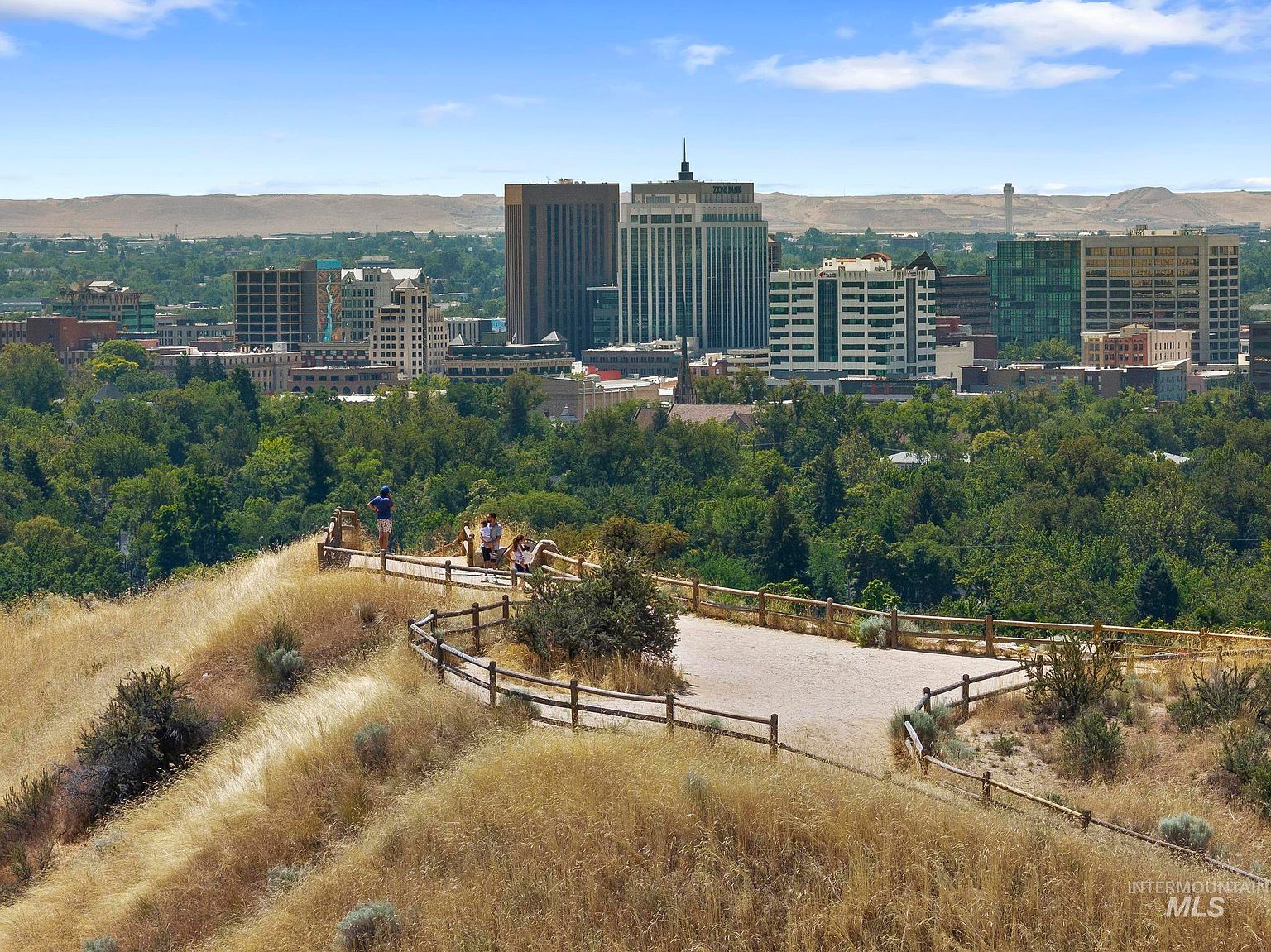 This elevated perspective captures a scenic overlook featuring a wooden railed path and a gravel viewing area where people are gathered. The foreground showcases dry, golden grasses typical of a hillside trail, while the background provides a panoramic view of a city skyline nestled against distant, arid mountains. The composition emphasizes the connection between natural recreational spaces and urban proximity.