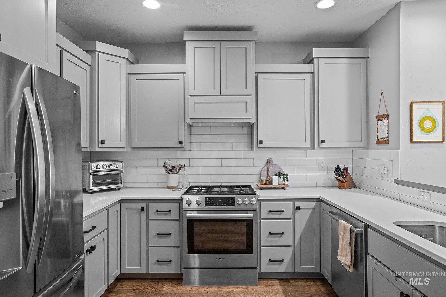 This modern kitchen features light gray cabinetry paired with white countertops and a classic white subway tile backsplash. The space is equipped with stainless steel appliances, including a gas range and a large refrigerator, creating a clean and functional aesthetic. The perspective is a straight-on, eye-level shot that highlights the symmetrical layout and the warm wood-toned flooring.