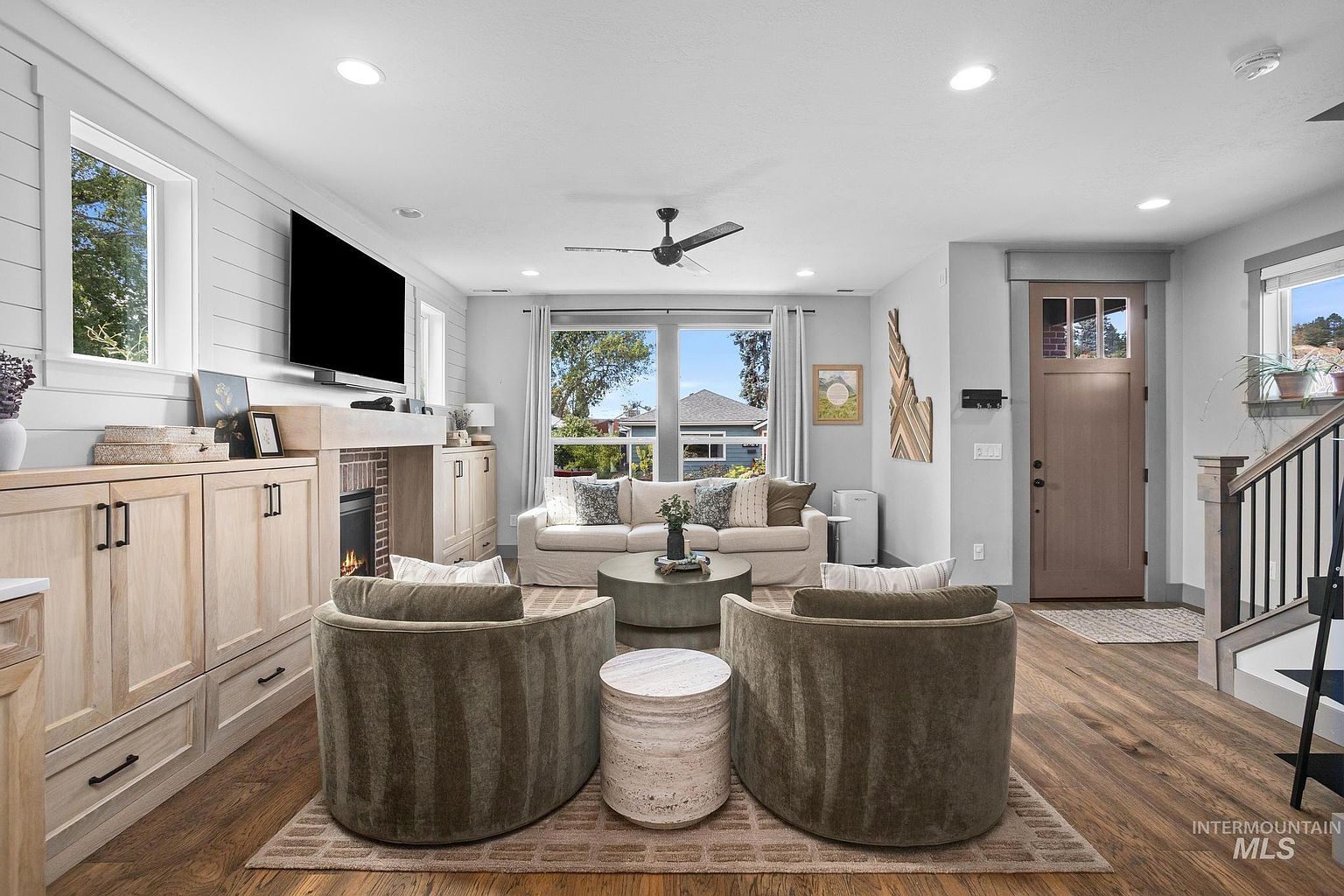 This bright and inviting living room features a neutral color palette with light wood cabinetry and warm-toned flooring. The space is anchored by a central fireplace with a mantel, flanked by built-in storage, and includes a comfortable sofa and two plush armchairs arranged around a circular coffee table. Large windows allow natural light to fill the room, creating an open and airy atmosphere perfect for relaxation.