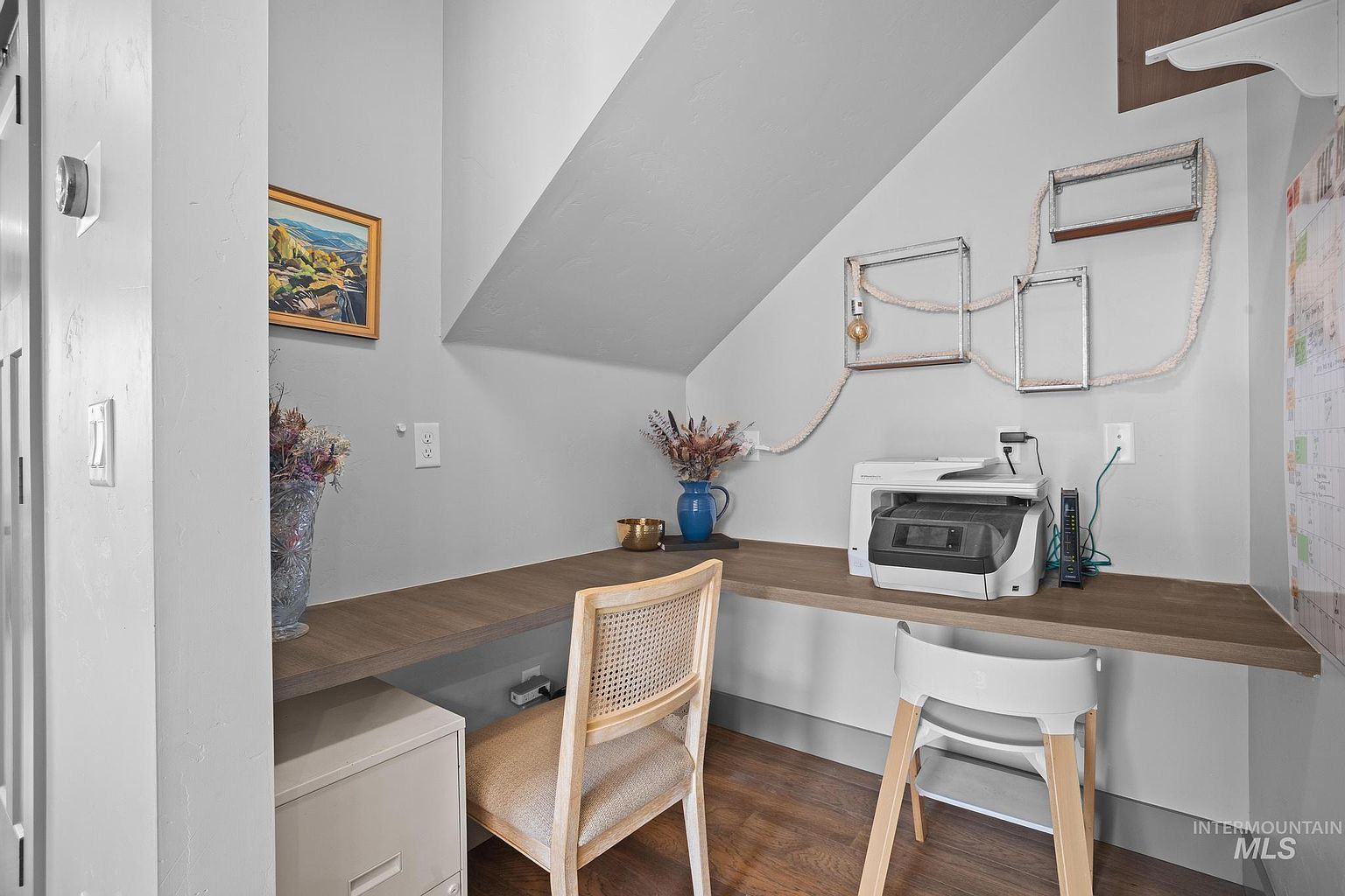 This cozy, tucked-away home office nook features a built-in desk with a wood-tone surface and a white filing cabinet underneath. The space is accented by a decorative wall display with geometric frames and a light-colored rope detail, alongside a small printer and a blue vase. The perspective is a straight-on shot of the workspace, highlighting the efficient use of an alcove area under a sloped ceiling.
