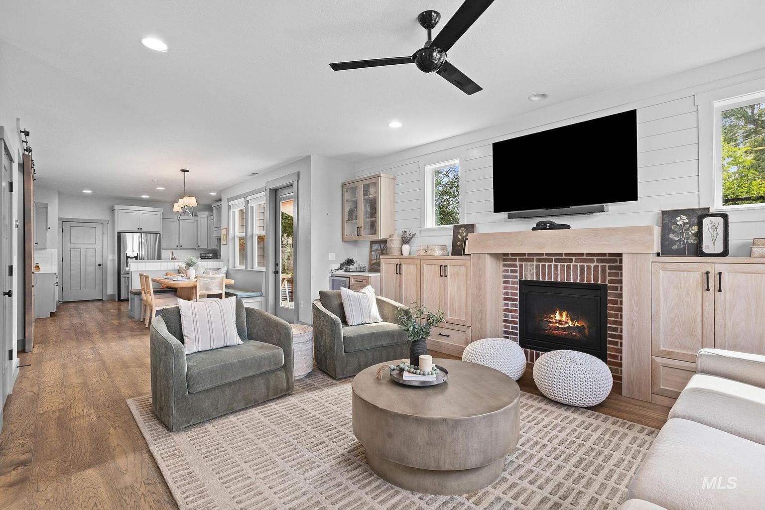 This open-concept living area features a cozy fireplace with a brick surround and a light wood mantel, flanked by custom cabinetry. Two plush armchairs face a central round coffee table, while a neutral area rug anchors the space beneath a modern ceiling fan. The room flows seamlessly into a bright dining area and kitchen, creating an inviting and cohesive living environment.