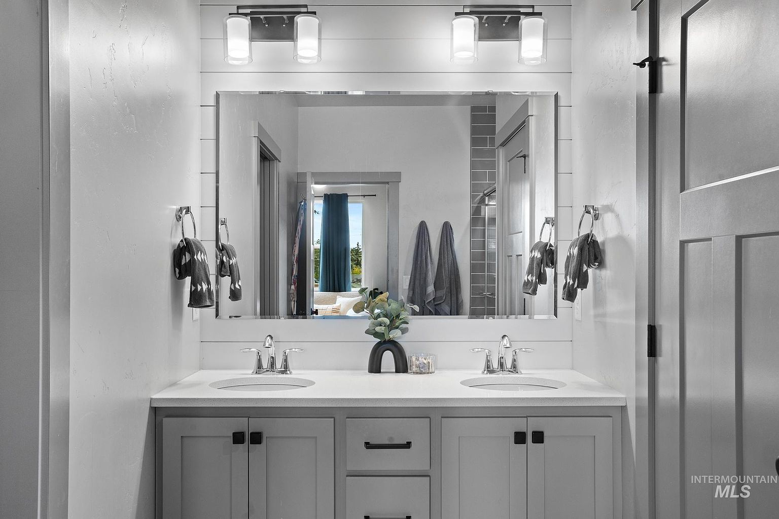 This modern double-vanity bathroom features a light gray cabinet base with dark hardware and a clean white countertop. A large, rectangular mirror is centered above the sinks, reflecting the shiplap wall detail and the doorway leading to an adjacent room. The space is illuminated by two contemporary wall-mounted light fixtures, creating a bright and polished aesthetic.