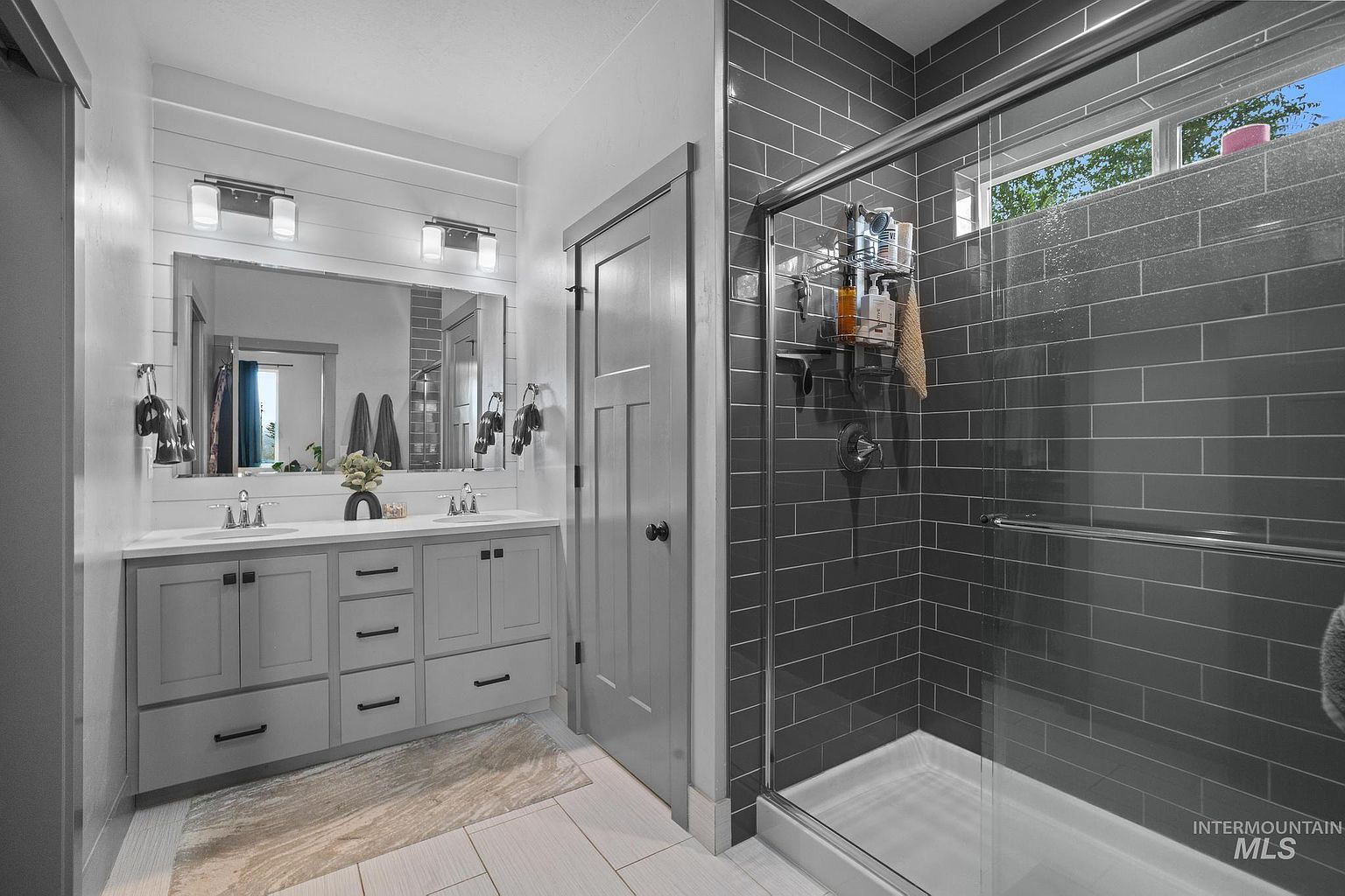 This modern bathroom features a double-sink vanity with gray cabinetry and a white countertop, set against a shiplap wall with a large mirror. To the right, a walk-in shower is enclosed by a glass door and finished with dark, rectangular subway-style wall tiles. The space is bright and clean, utilizing a neutral color palette and contemporary fixtures to create a polished, functional aesthetic.