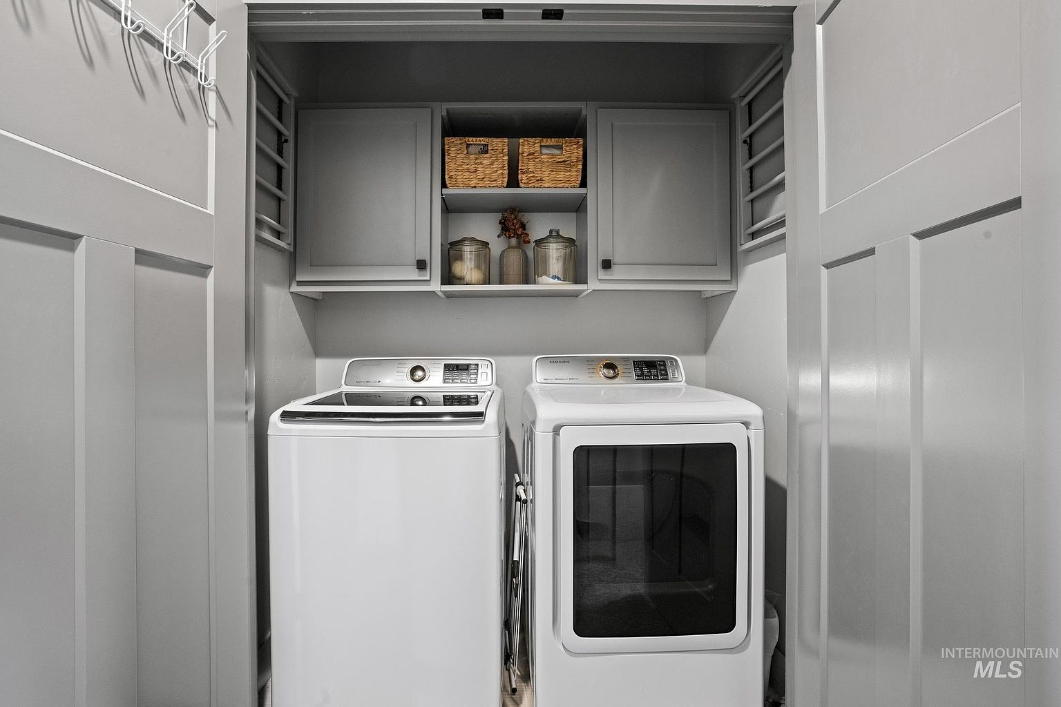 This compact and organized laundry room features a side-by-side washer and dryer set tucked into a dedicated closet space. Above the appliances, custom gray cabinetry provides convenient storage, complemented by an open shelf holding decorative baskets and glass canisters. The neutral color palette and clean lines create a functional and tidy utility area.