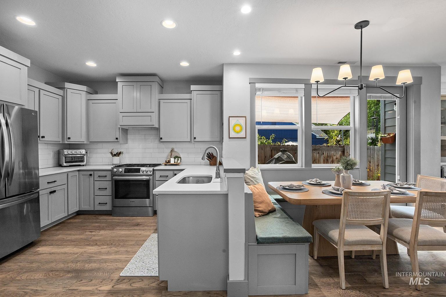 This bright, modern kitchen features light gray cabinetry, a white subway tile backsplash, and stainless steel appliances. A peninsula with a sink separates the cooking area from a cozy dining nook, which includes a built-in bench seat and a wooden table with four chairs. The space is illuminated by recessed lighting and a contemporary chandelier, creating an inviting and functional open-concept layout.