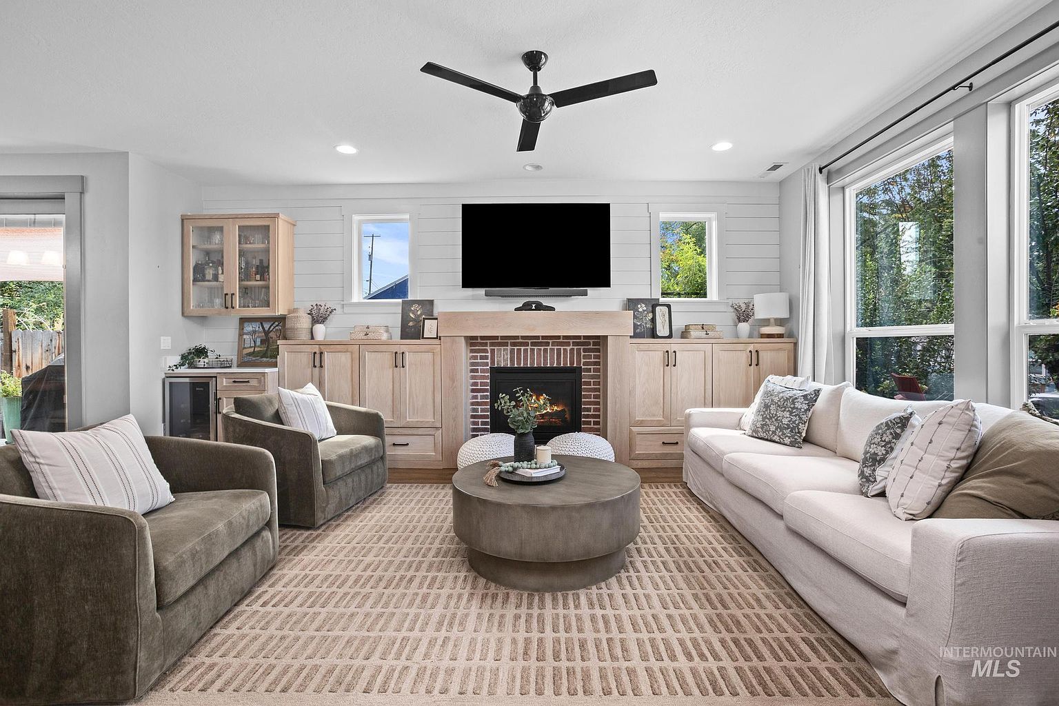This bright and inviting living room features a central fireplace with a brick surround and a light wood mantel, flanked by custom cabinetry. The space is furnished with a large, light-colored sofa and two comfortable armchairs arranged around a round, neutral-toned coffee table on a textured area rug. Large windows on the right side of the room allow for ample natural light, creating an airy and welcoming atmosphere.