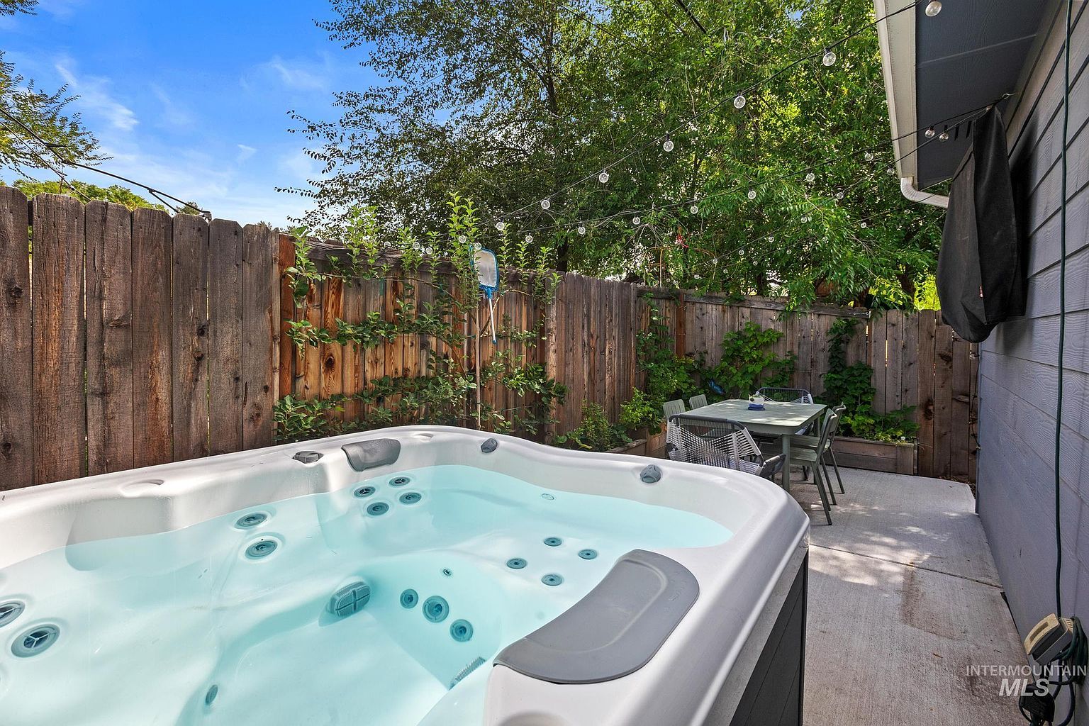 This inviting outdoor living space features a modern hot tub positioned on a concrete patio, perfect for relaxation. Adjacent to the spa, a dining set sits under a string of decorative globe lights, creating a cozy atmosphere against the backdrop of a wooden privacy fence and lush greenery. The perspective captures a private, well-maintained backyard retreat that blends functional leisure space with natural elements.