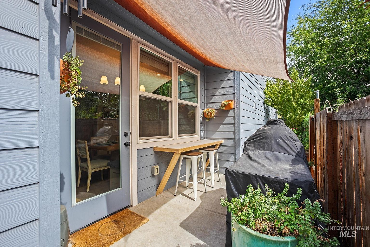 This cozy outdoor patio space features a light wood bar table with two stools positioned against the exterior wall, perfect for casual dining. A large, covered grill sits to the right, while a shade sail provides overhead protection from the sun. The area is accented with hanging planters and a large potted plant, creating a welcoming and functional outdoor living environment.
